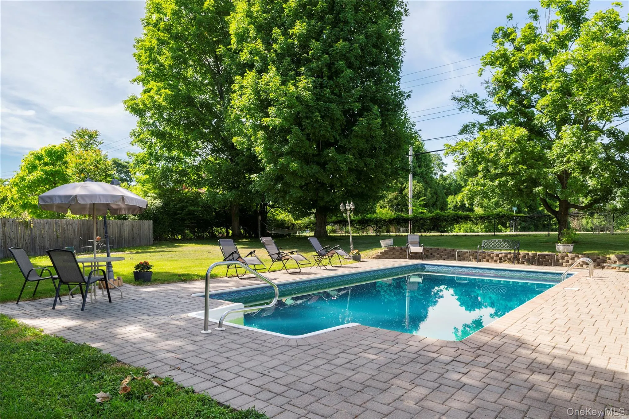 Outdoor swimming pool surrounded by a paver patio and expansive green lawn, featuring mature shade trees and a privacy fence Outdoor swimming pool surrounded by a paver patio and expansive green lawn, featuring mature shade trees and a privacy fence