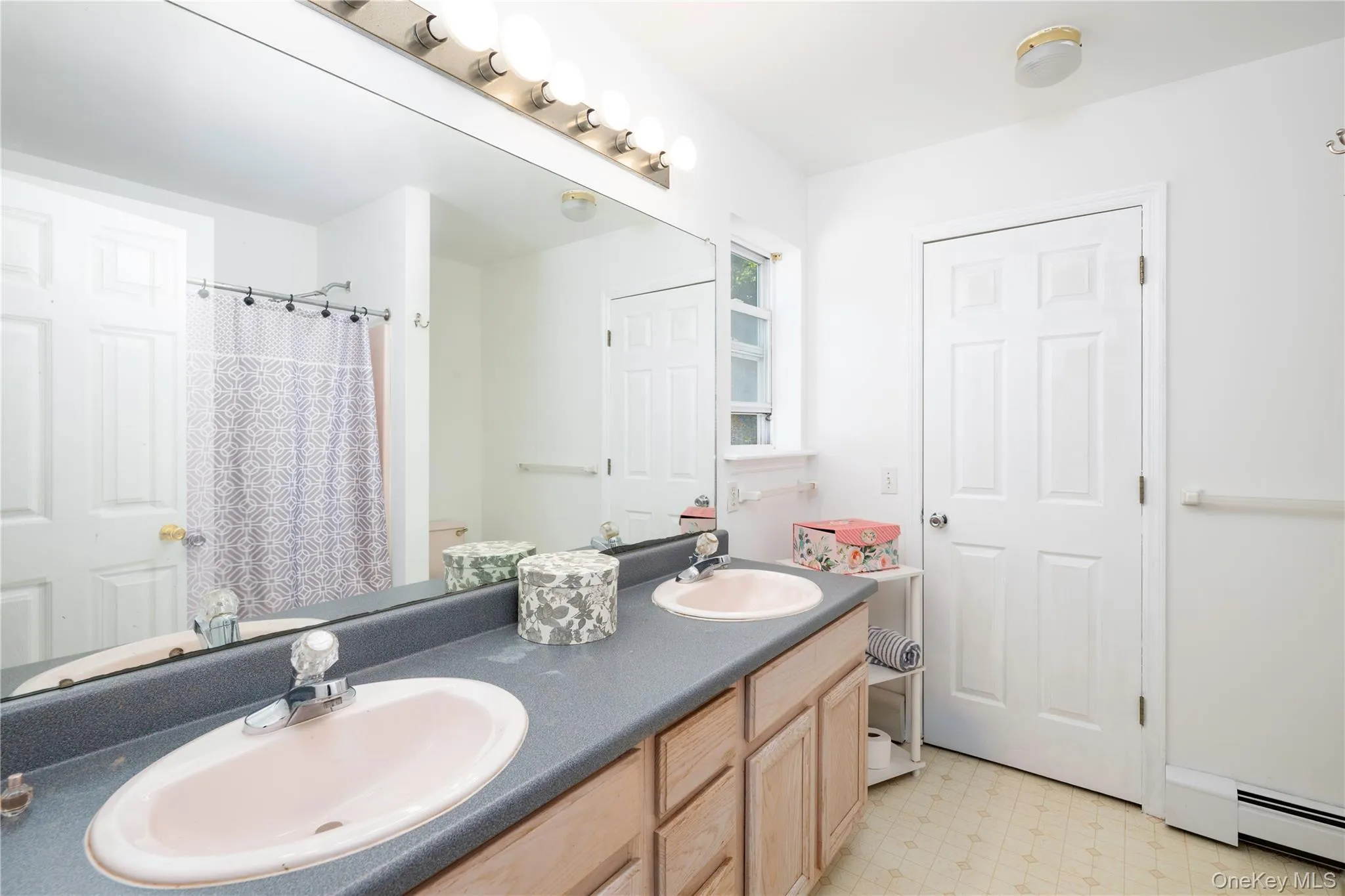 Bathroom featuring a dual-sink vanity with light wood-finish cabinetry, a large wall-mounted mirror, and overhead strip lighting Bathroom featuring a dual-sink vanity with light wood-finish cabinetry, a large wall-mounted mirror, and overhead strip lighting