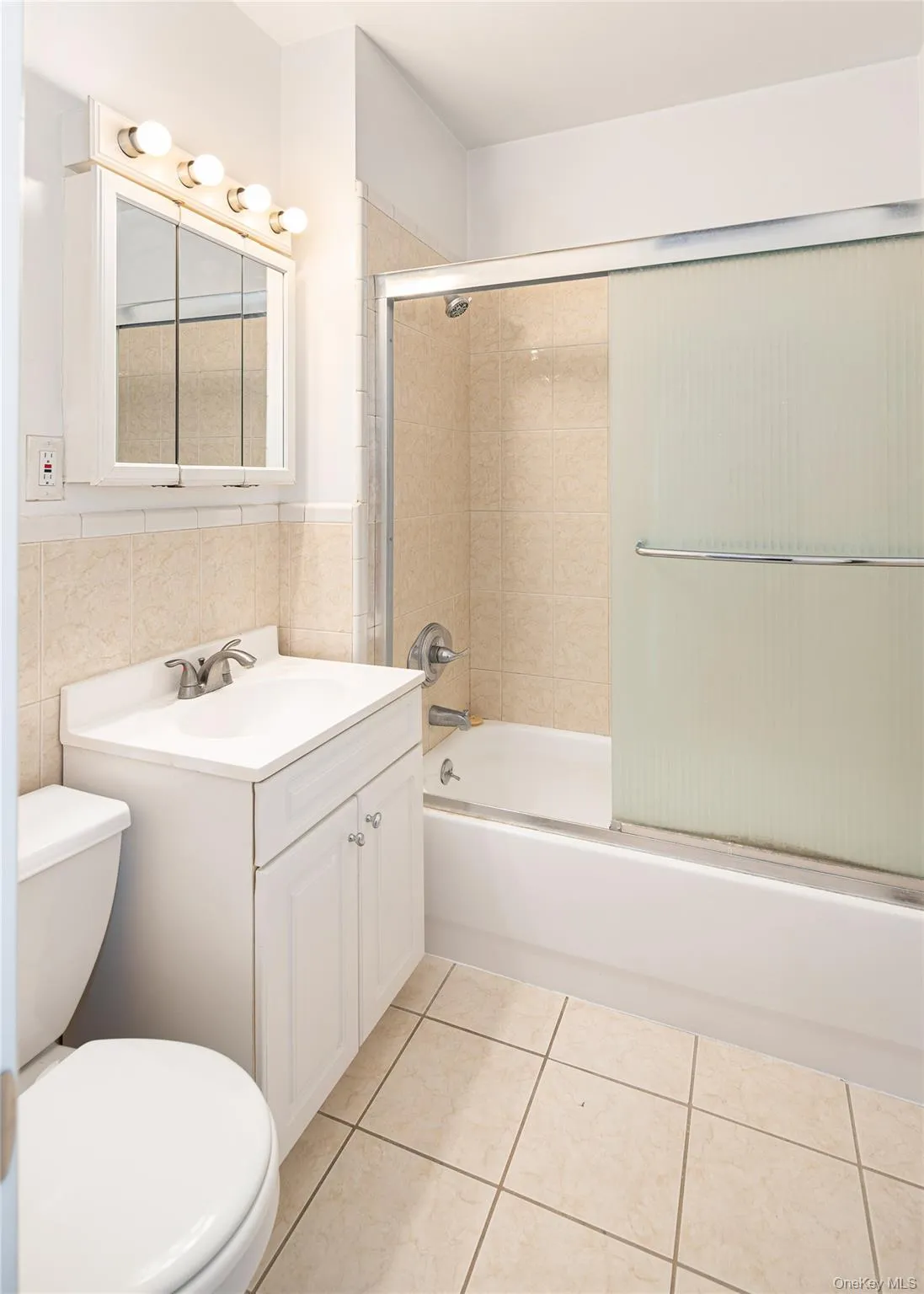 Bathroom featuring vanity, bath / shower combo with glass door, tile walls, light tile patterned flooring, and a wainscoted wall Bathroom featuring vanity, bath / shower combo with glass door, tile walls, light tile patterned flooring, and a wainscoted wall