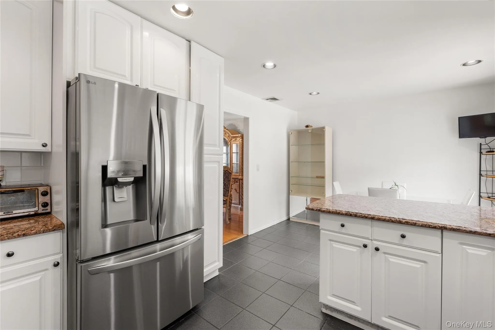 Modern kitchen featuring white cabinetry, granite countertops, a stainless steel refrigerator, recessed lighting, and dark tile flooring Modern kitchen featuring white cabinetry, granite countertops, a stainless steel refrigerator, recessed lighting, and dark tile flooring