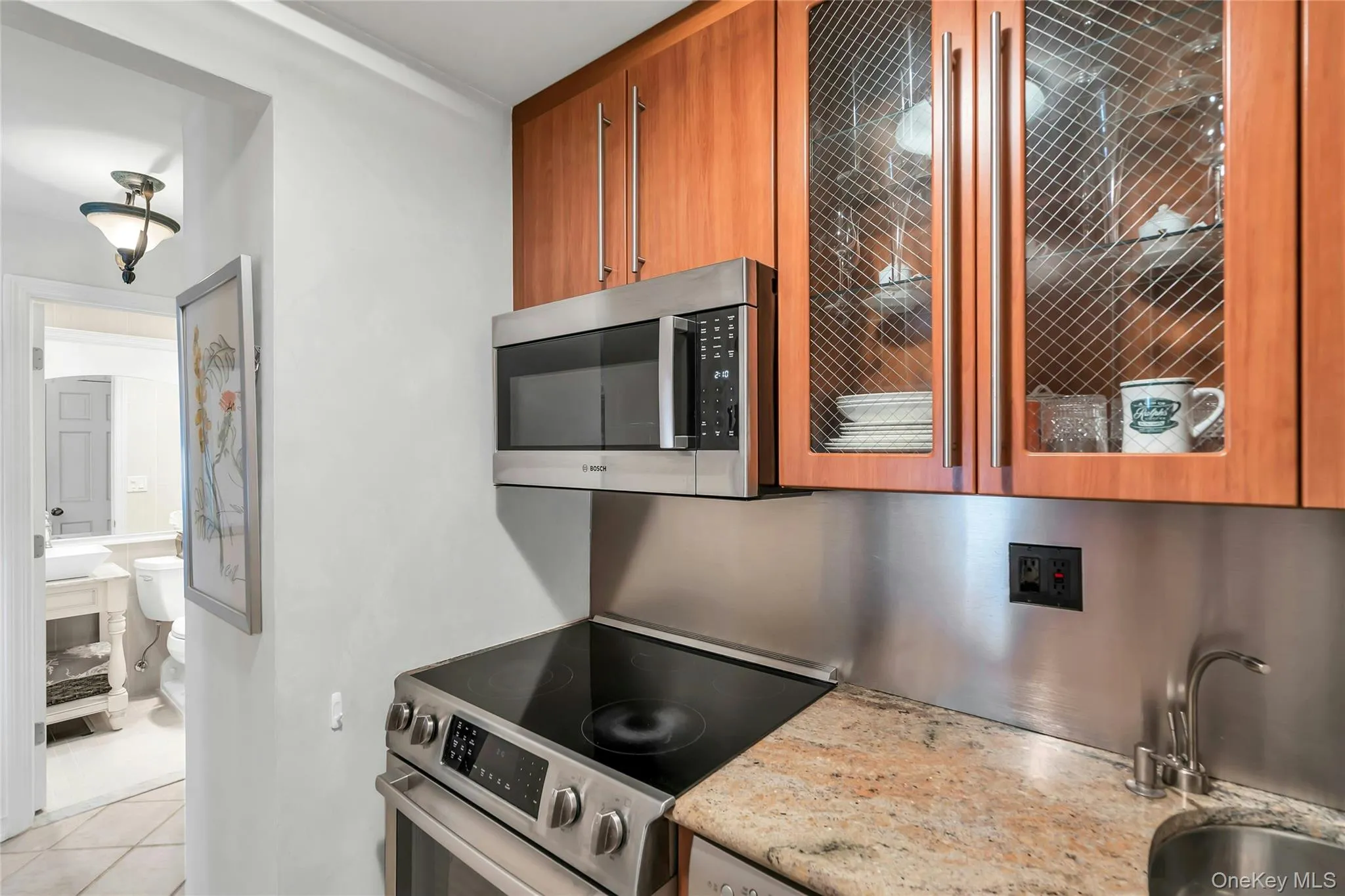 Kitchen area featuring stainless steel Bosche and Perlick under the Cabinet, Refrigerator appliances, wood-finish cabinetry with decorative glass inserts, stone-finish countertops, and a stainless steel back backsplash Kitchen area featuring stainless steel Bosche and Perlick under the Cabinet, Refrigerator appliances, wood-finish cabinetry with decorative glass inserts, stone-finish countertops, and a stainless steel back backsplash