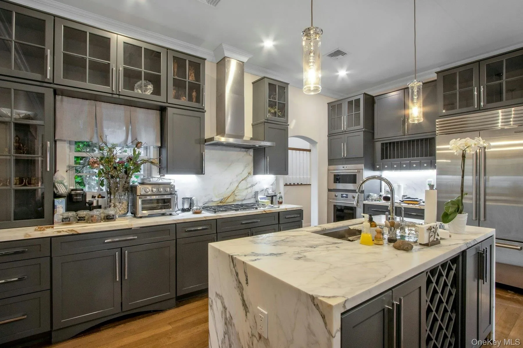 Kitchen featuring wall chimney exhaust hood, stainless steel appliances, light stone counters, an island with sink, and decorative light fixtures Kitchen featuring wall chimney exhaust hood, stainless steel appliances, light stone counters, an island with sink, and decorative light fixtures