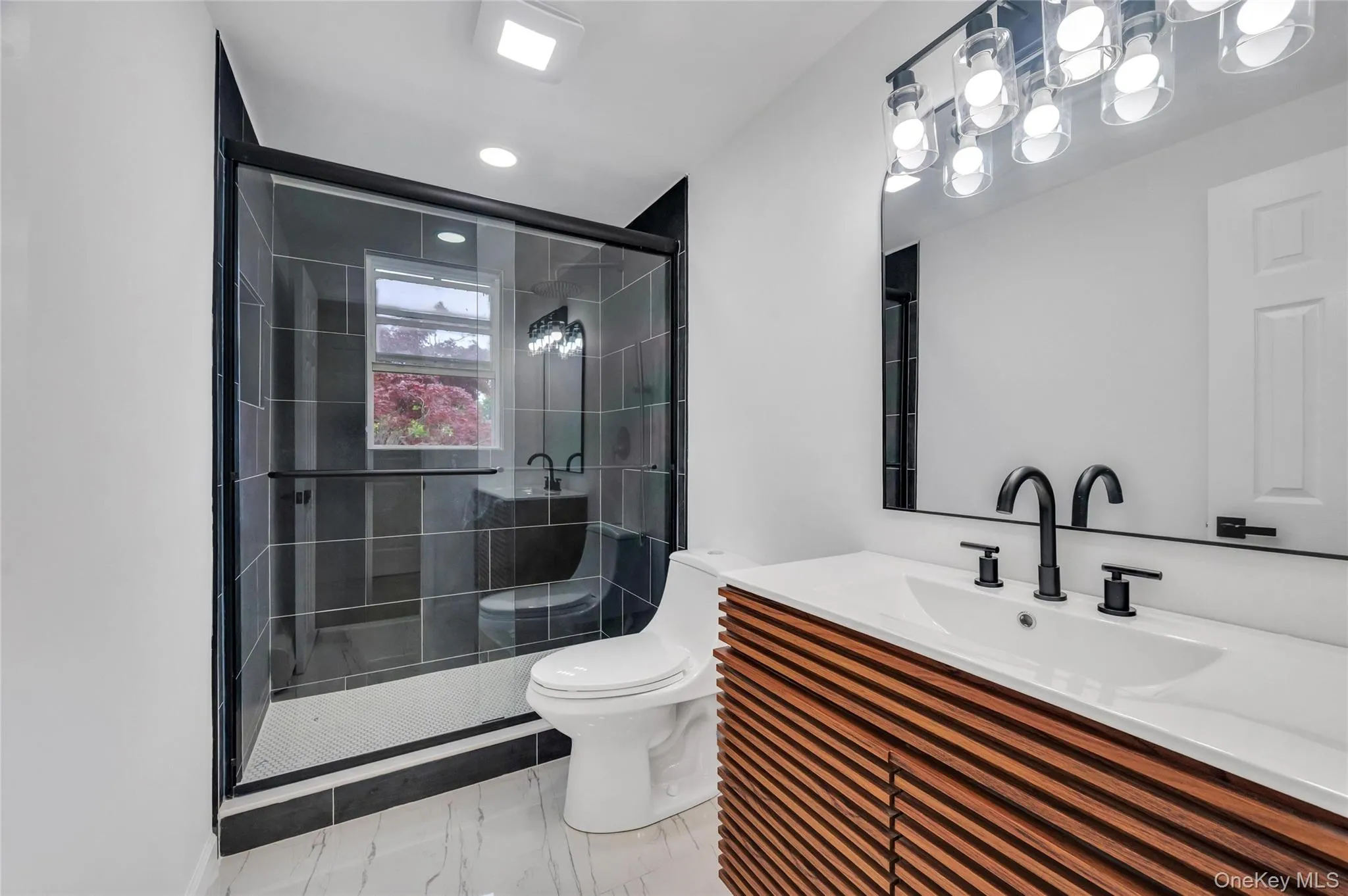 Modern bathroom featuring a glass-enclosed shower with dark tile surround, a slatted wood-finish vanity with an integrated sink, matte black fixtures, and a multi-bulb vanity light Modern bathroom featuring a glass-enclosed shower with dark tile surround, a slatted wood-finish vanity with an integrated sink, matte black fixtures, and a multi-bulb vanity light