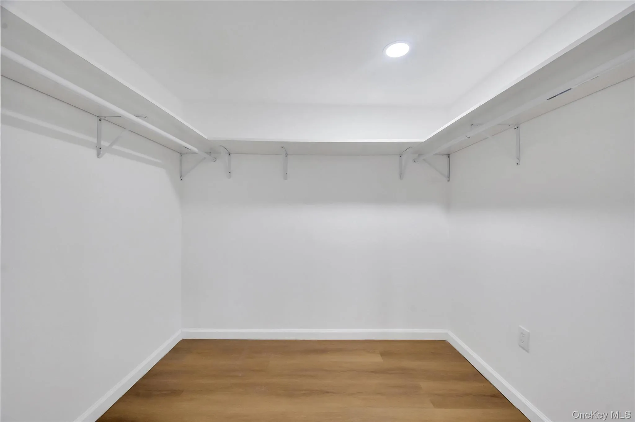 Walk-in closet featuring wood-finish flooring, white baseboards, recessed lighting, and wrap-around shelving with hanging rods Walk-in closet featuring wood-finish flooring, white baseboards, recessed lighting, and wrap-around shelving with hanging rods