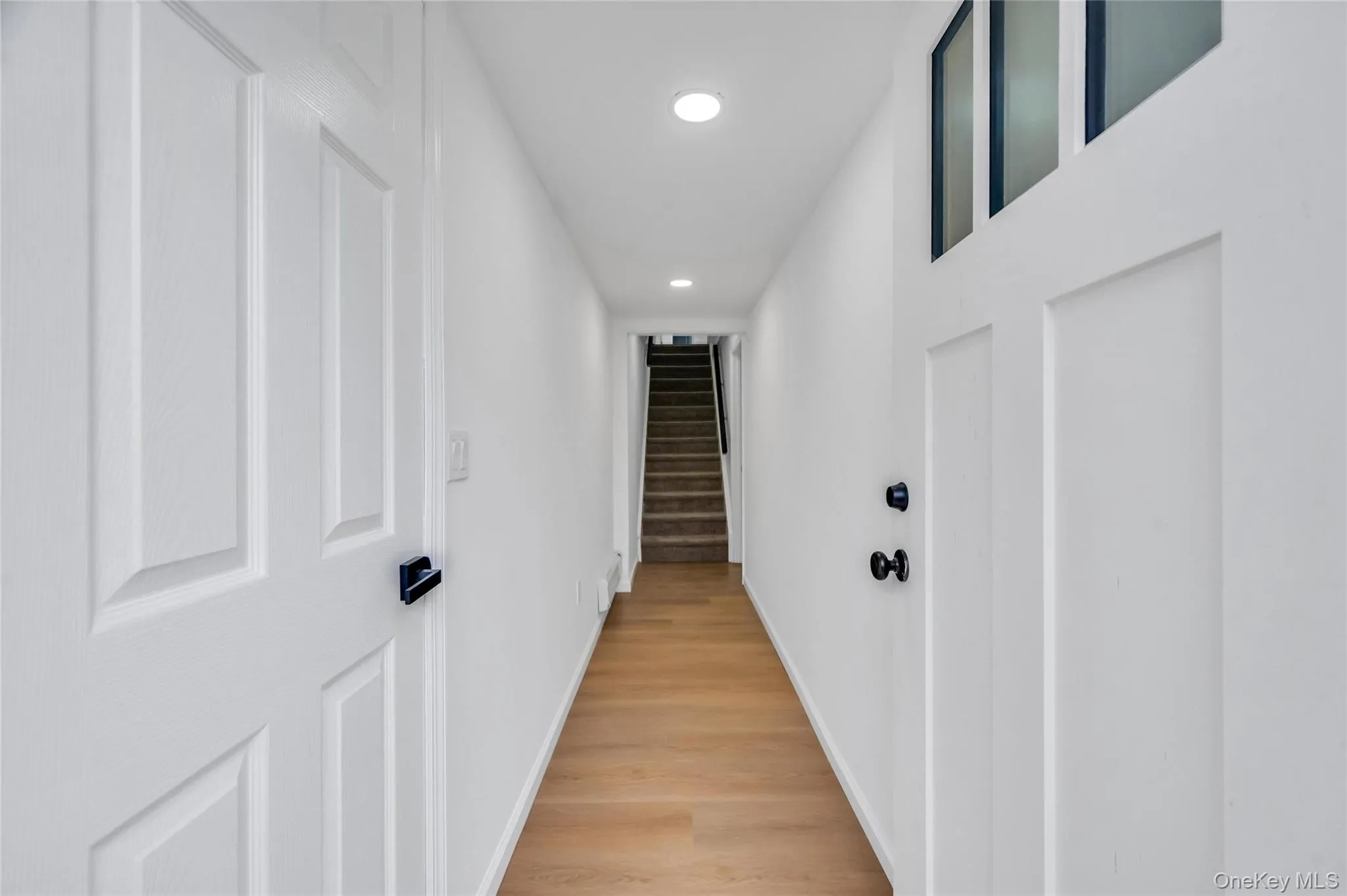Hallway featuring wood-finish flooring, recessed lighting, and white paneled doors with black hardware Hallway featuring wood-finish flooring, recessed lighting, and white paneled doors with black hardware