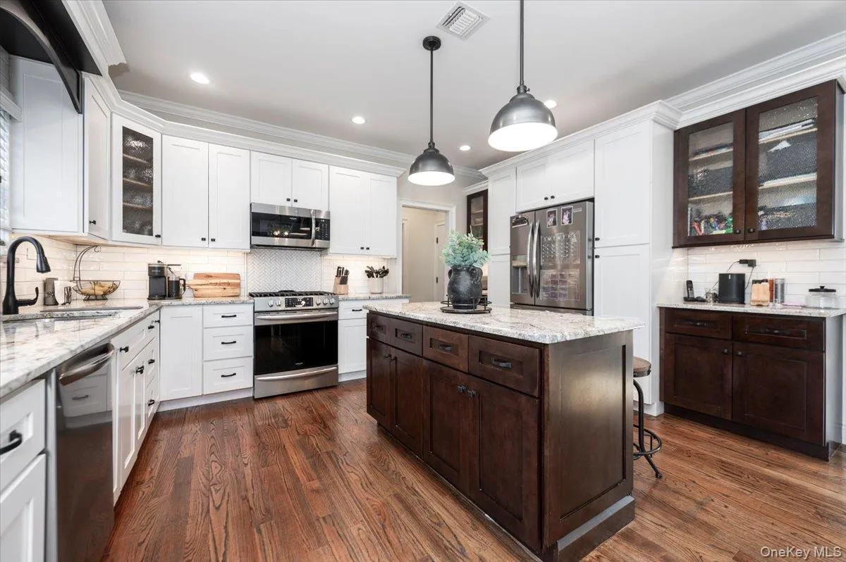 Kitchen featuring wood-finish flooring, a central island with a light-toned countertop, white perimeter cabinetry, stainless steel appliances, and a subway tile backsplash Kitchen featuring wood-finish flooring, a central island with a light-toned countertop, white perimeter cabinetry, stainless steel appliances, and a subway tile backsplash