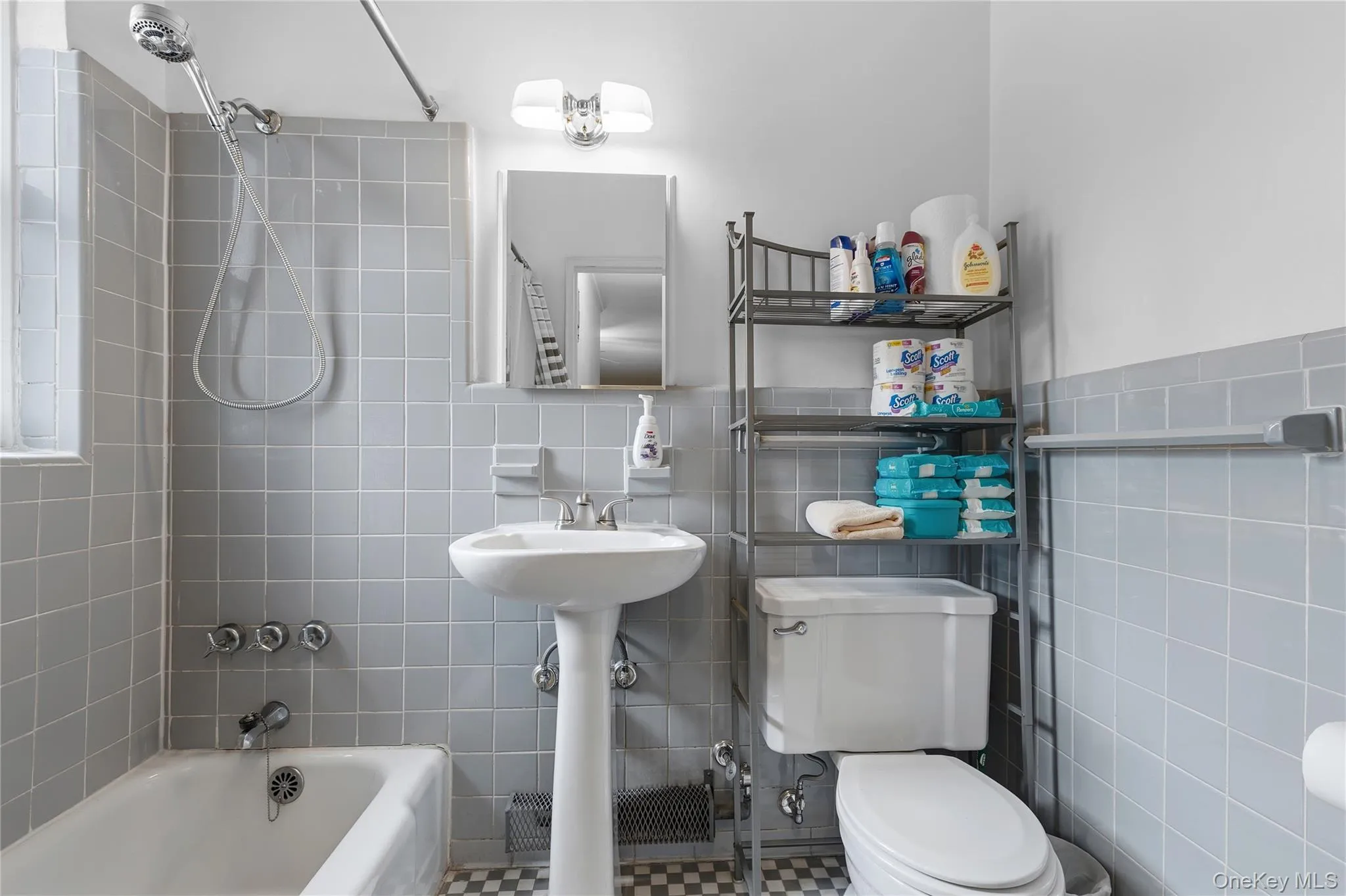 Bathroom featuring gray square wall tiling, a white pedestal sink, a white bathtub with a shower attachment, and checkerboard floor tiling Bathroom featuring gray square wall tiling, a white pedestal sink, a white bathtub with a shower attachment, and checkerboard floor tiling