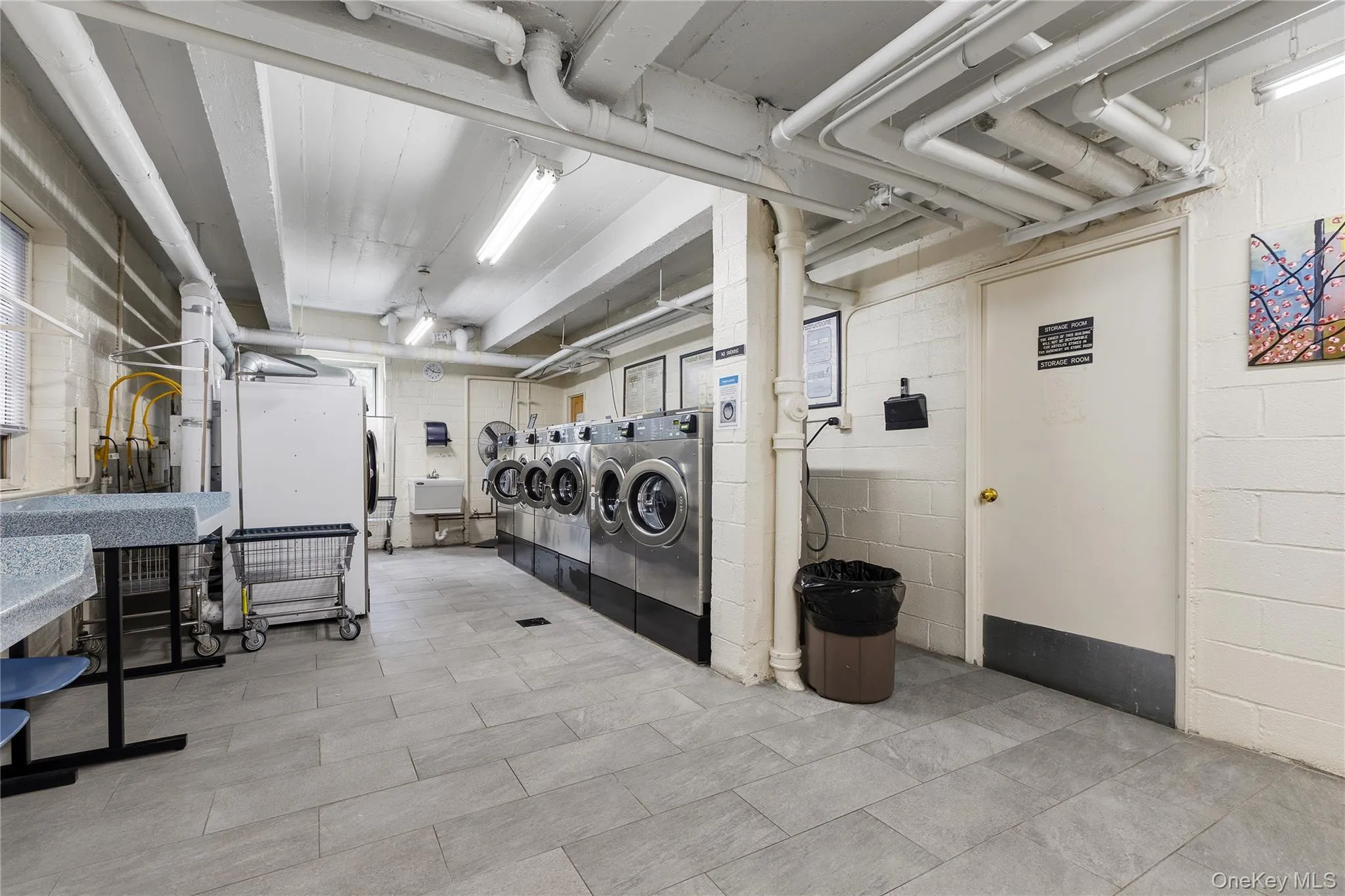 Well-lit laundry facility featuring multiple front-loading washing machines, dedicated drying machines, and a utility sink Well-lit laundry facility featuring multiple front-loading washing machines, dedicated drying machines, and a utility sink