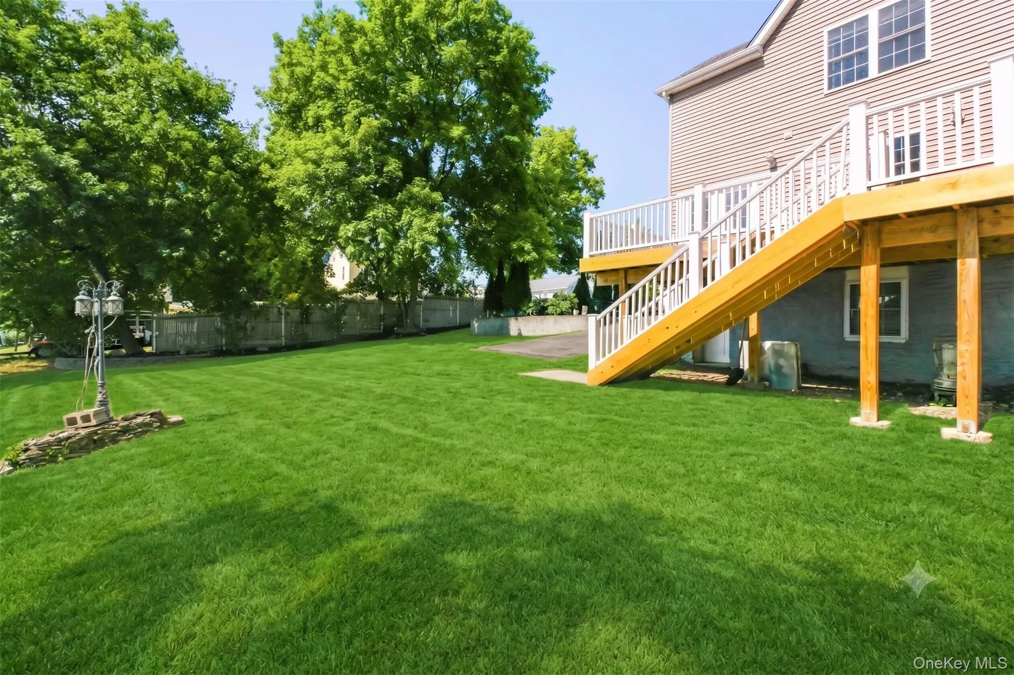 Virtually Staged. Rear exterior view highlighting the elevated deck, staircase, fenced side area, driveway access, and generous backyard space with mature greenery. Virtually Staged. Rear exterior view highlighting the elevated deck, staircase, fenced side area, driveway access, and generous backyard space with mature greenery.
