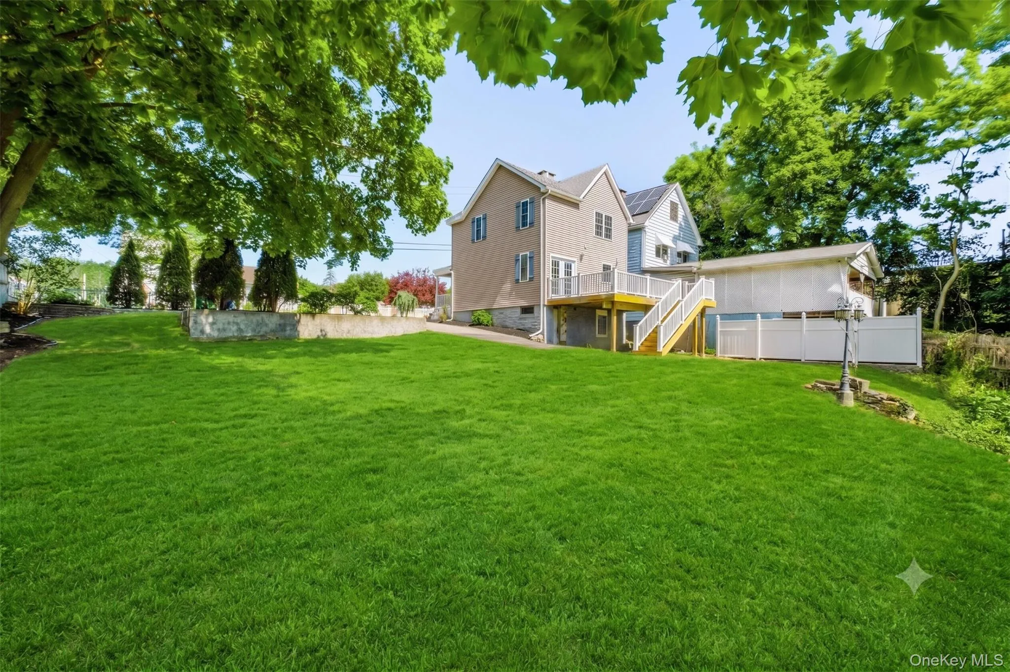 Virtually Staged. Expansive rear yard featuring a large lawn, mature landscaping, rear deck with staircase access, driveway area, and surrounding greenery that adds privacy and charm. Virtually Staged. Expansive rear yard featuring a large lawn, mature landscaping, rear deck with staircase access, driveway area, and surrounding greenery that adds privacy and charm.