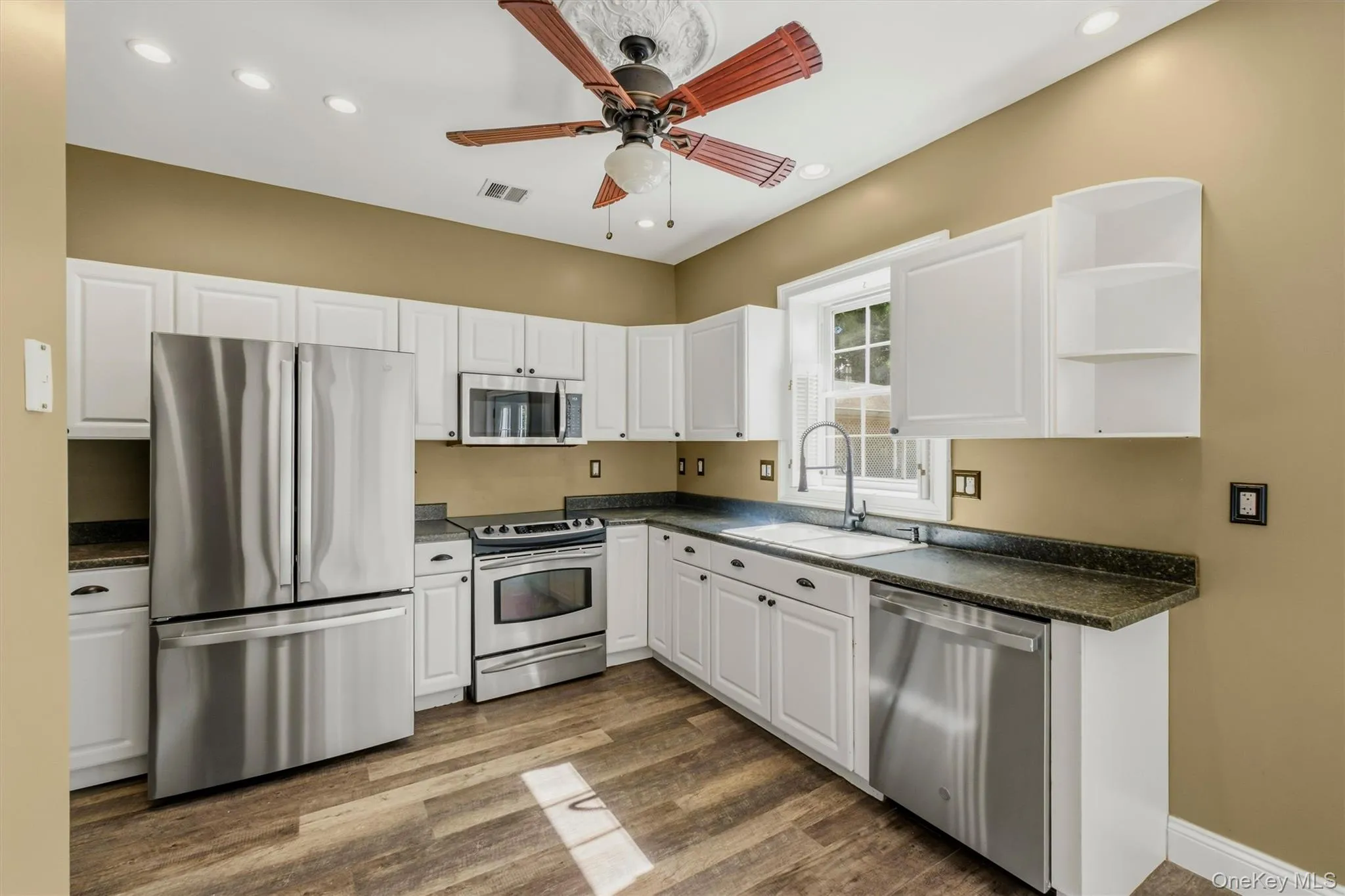 Spacious kitchen featuring white cabinetry, stainless steel appliances, dark countertops, wood-look flooring, recessed lighting, and a ceiling fan, offering a functional layout with plenty of room to move around. Spacious kitchen featuring white cabinetry, stainless steel appliances, dark countertops, wood-look flooring, recessed lighting, and a ceiling fan, offering a functional layout with plenty of room to move around.