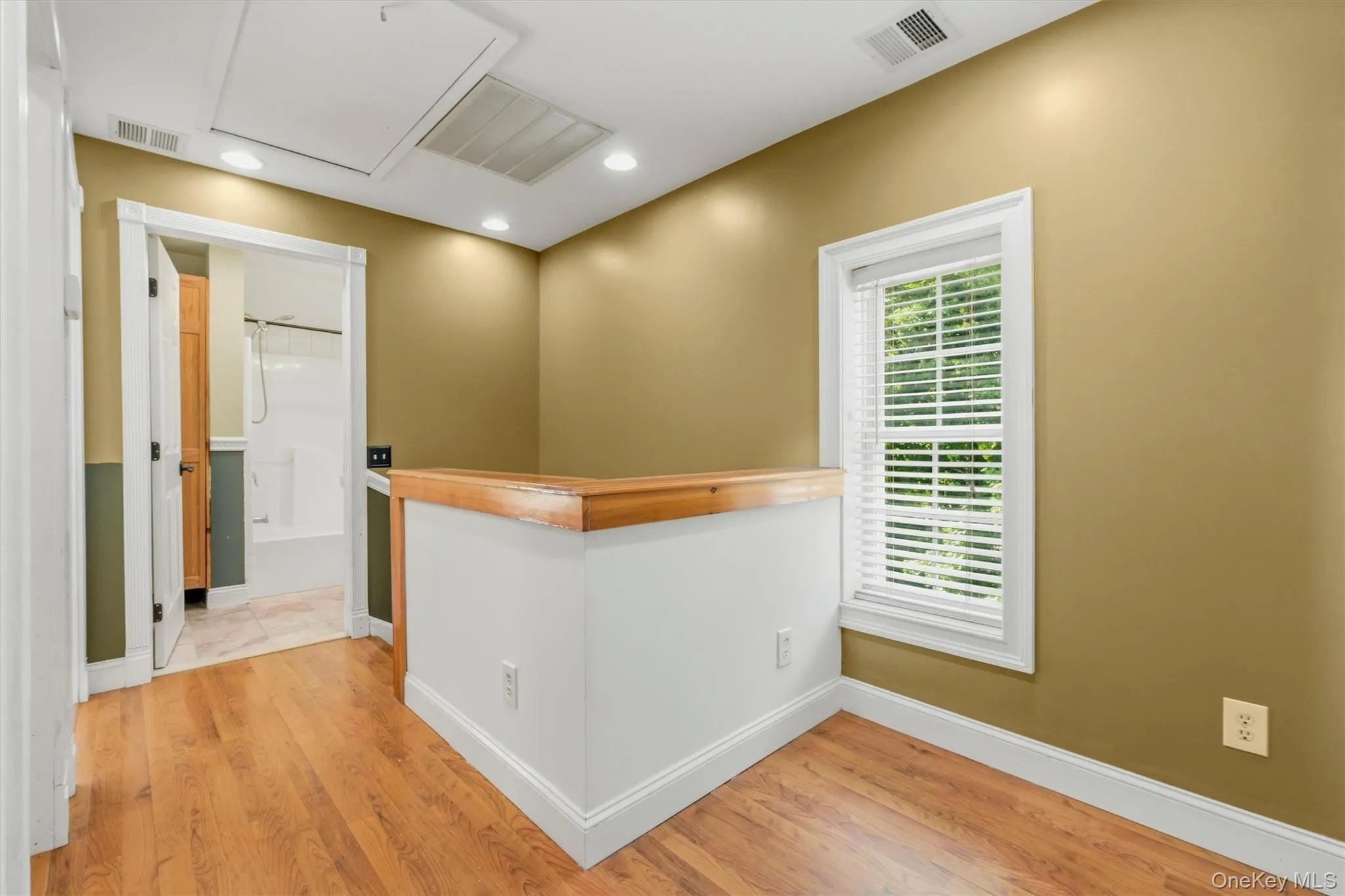 Upper-level hallway featuring warm wood-style flooring, recessed lighting, white trim, a bright window, and direct access to the full bathroom, creating a functional transition space between rooms. Upper-level hallway featuring warm wood-style flooring, recessed lighting, white trim, a bright window, and direct access to the full bathroom, creating a functional transition space between rooms.