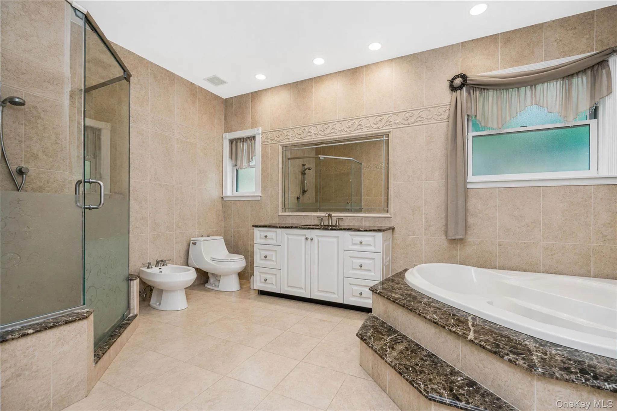 Spacious bathroom featuring a glass-enclosed shower, built-in soaking tub with stone surround, a white vanity with double sinks and stone countertop, and recessed lighting Spacious bathroom featuring a glass-enclosed shower, built-in soaking tub with stone surround, a white vanity with double sinks and stone countertop, and recessed lighting