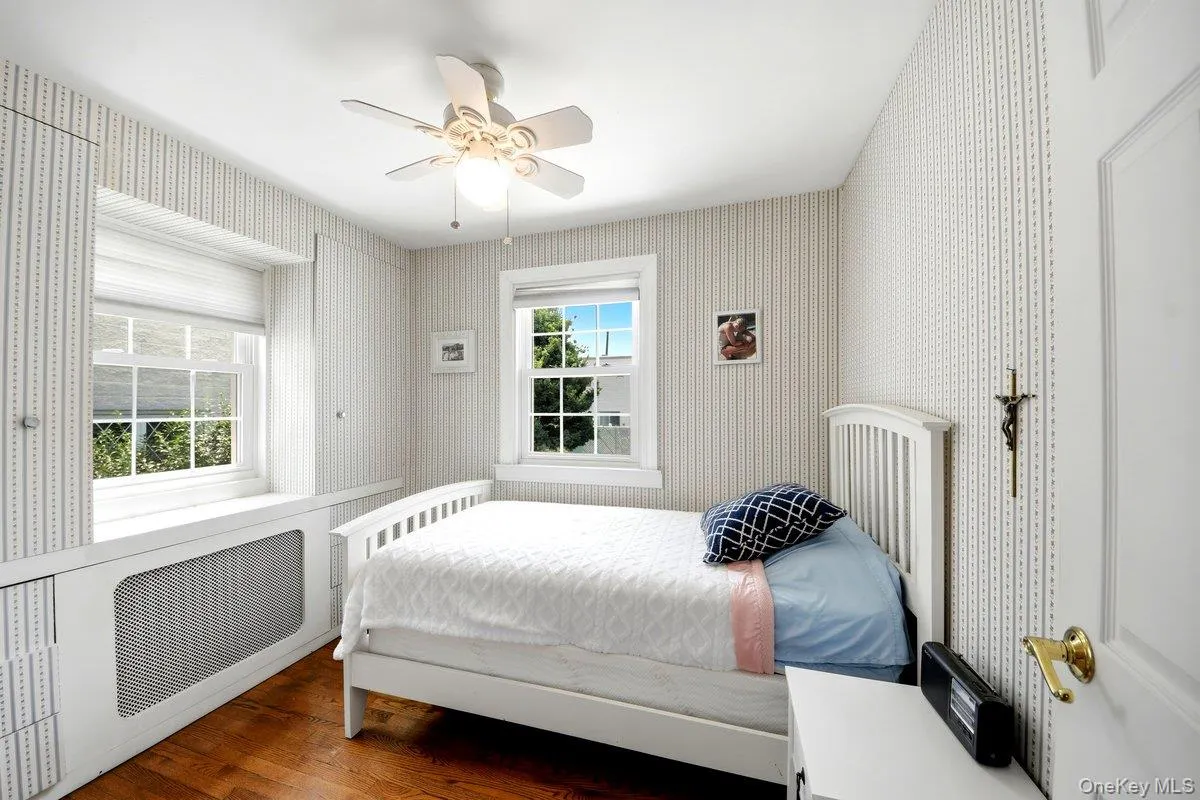 Hardwood flooring grounds this first floor bedroom, featuring two windows with white trim, a built-in radiator cover Hardwood flooring grounds this first floor bedroom, featuring two windows with white trim, a built-in radiator cover