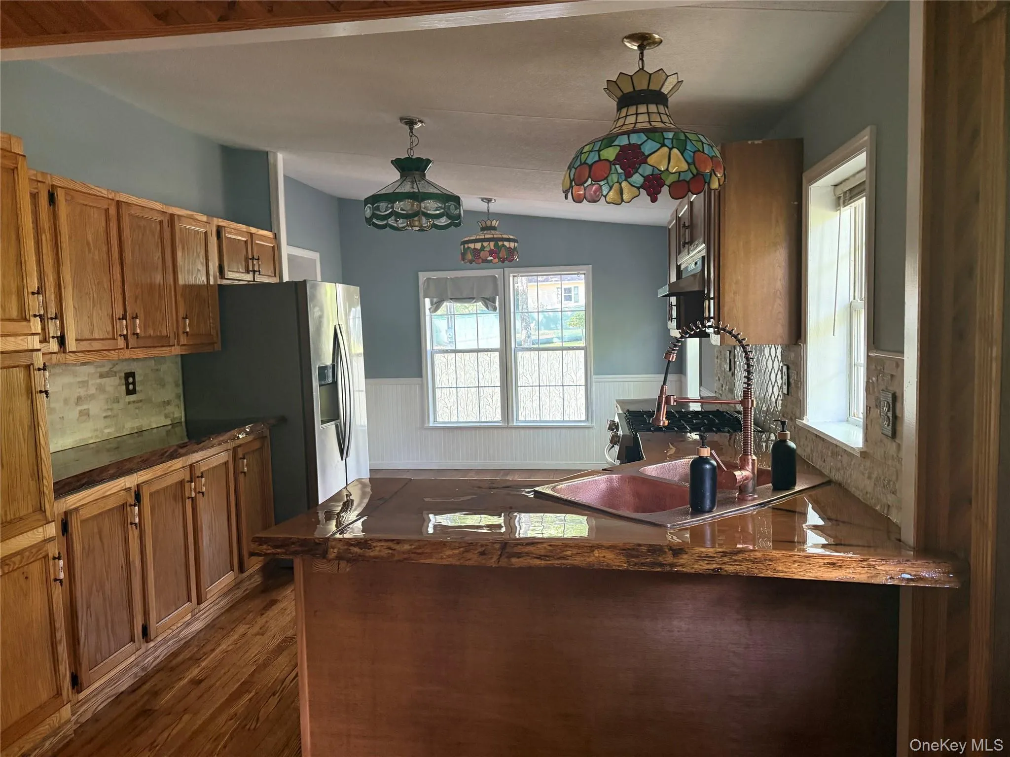 Kitchen with wainscoting, brown cabinetry, a peninsula, lofted ceiling, and dark wood-style floors Kitchen with wainscoting, brown cabinetry, a peninsula, lofted ceiling, and dark wood-style floors