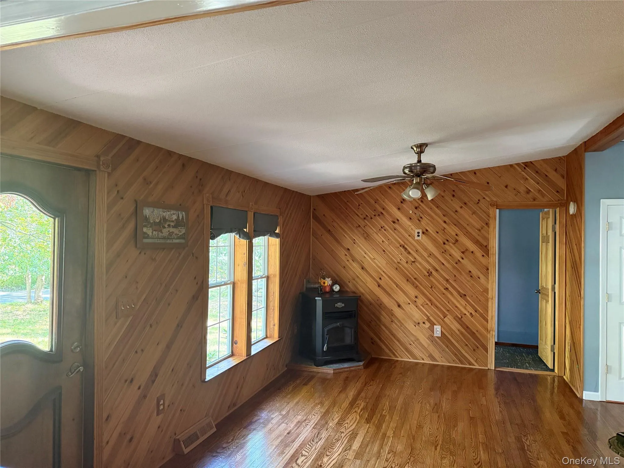 Unfurnished living room with wooden walls, wood finished floors, a wood stove, and ceiling fan Unfurnished living room with wooden walls, wood finished floors, a wood stove, and ceiling fan