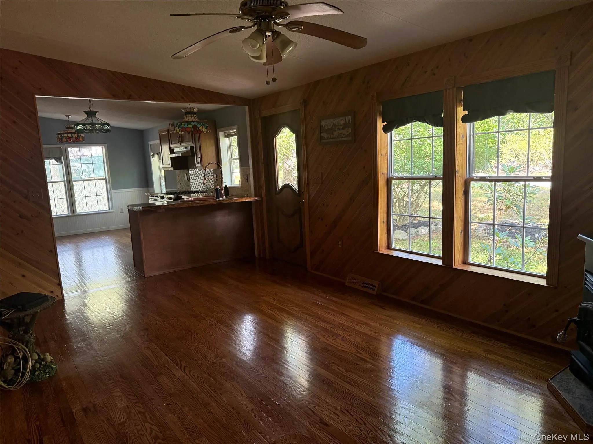 Unfurnished living room featuring wooden walls, dark wood-style flooring, ceiling fan, a wood stove, and wainscoting Unfurnished living room featuring wooden walls, dark wood-style flooring, ceiling fan, a wood stove, and wainscoting