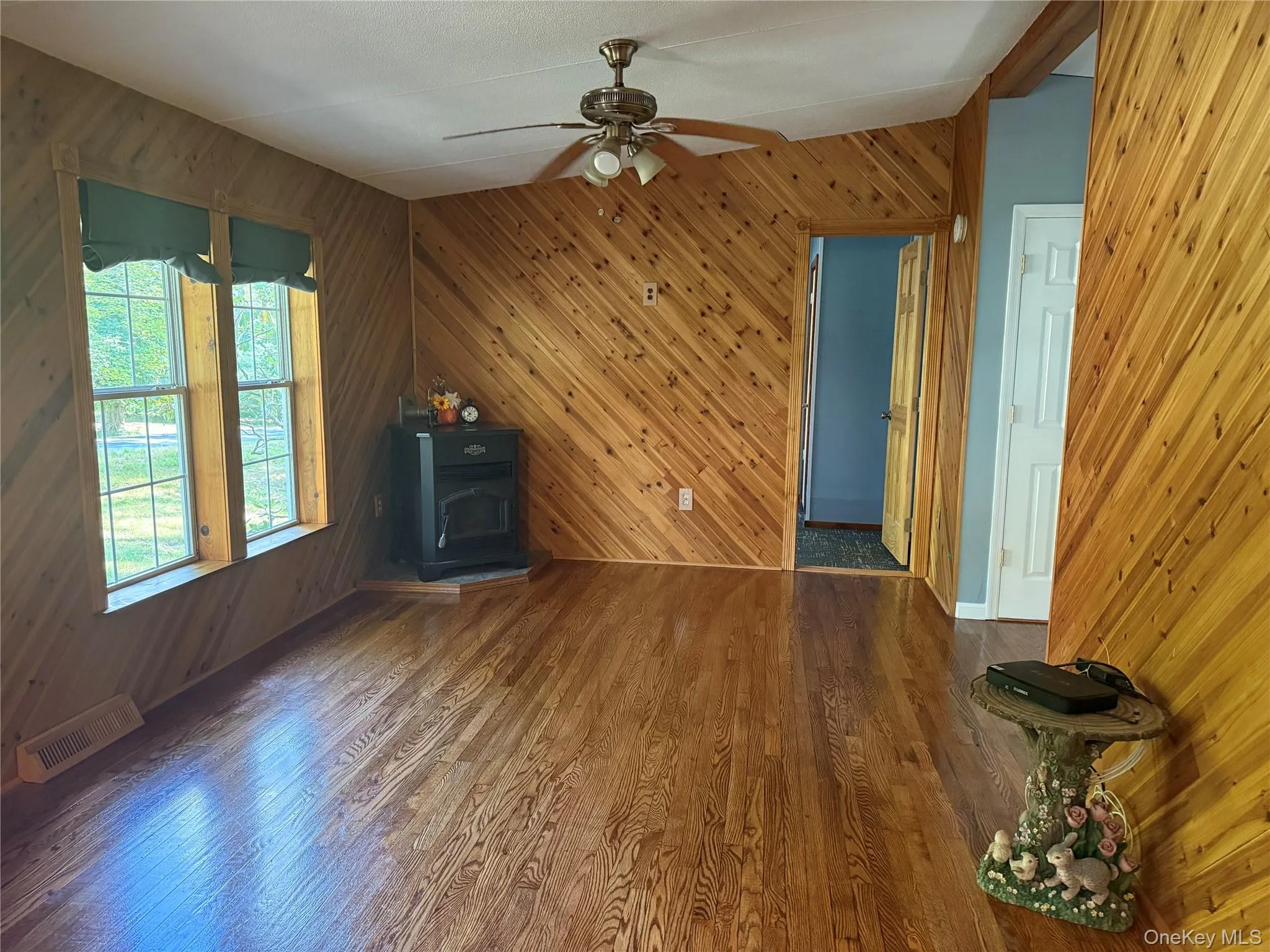 Unfurnished living room with wooden walls, wood finished floors, a wood stove, and a ceiling fan Unfurnished living room with wooden walls, wood finished floors, a wood stove, and a ceiling fan