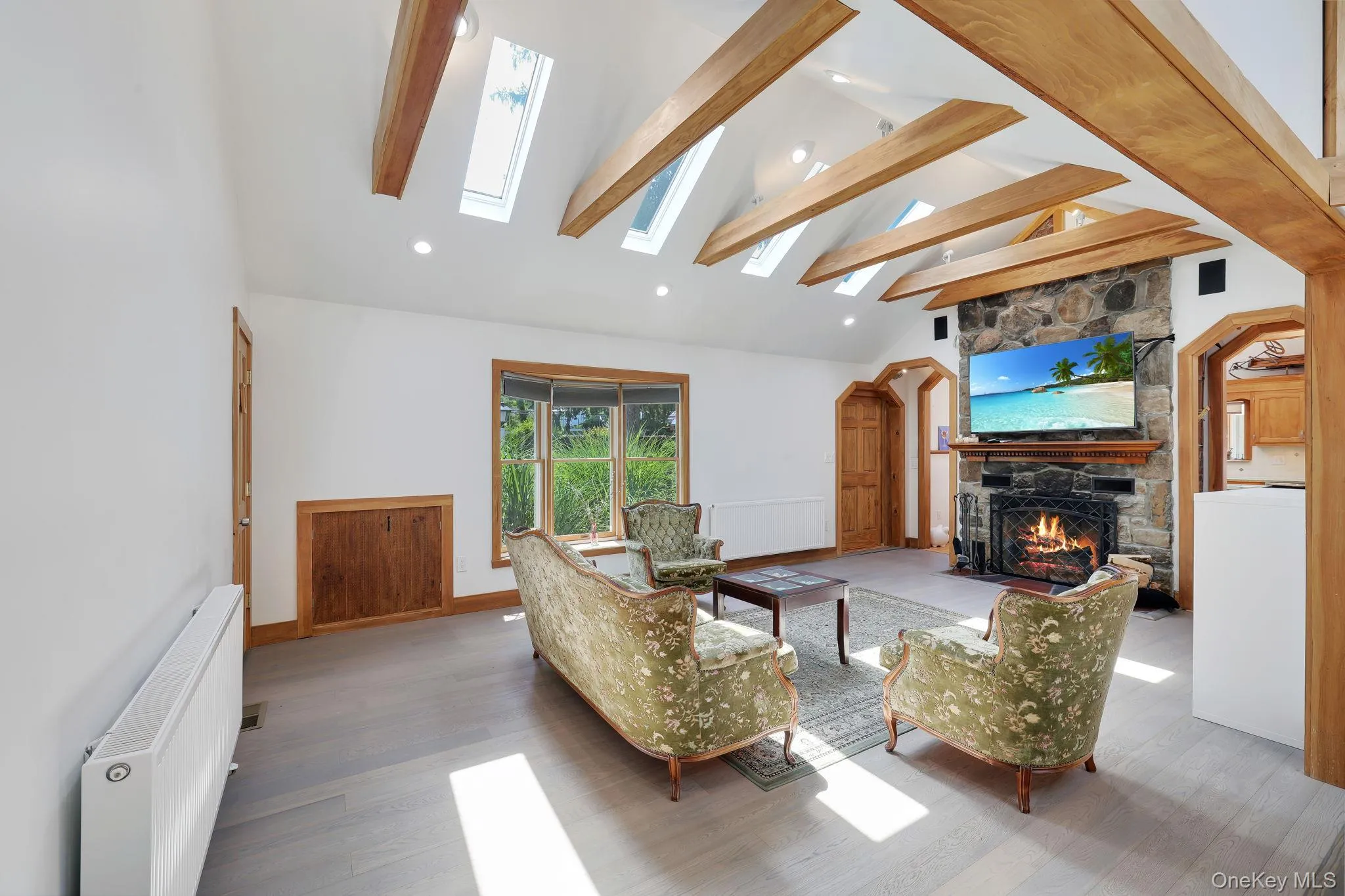 Spacious living room featuring a stone fireplace, a vaulted ceiling with exposed wood beams, multiple skylights, and light-toned wood-finish flooring. Spacious living room featuring a stone fireplace, a vaulted ceiling with exposed wood beams, multiple skylights, and light-toned wood-finish flooring.