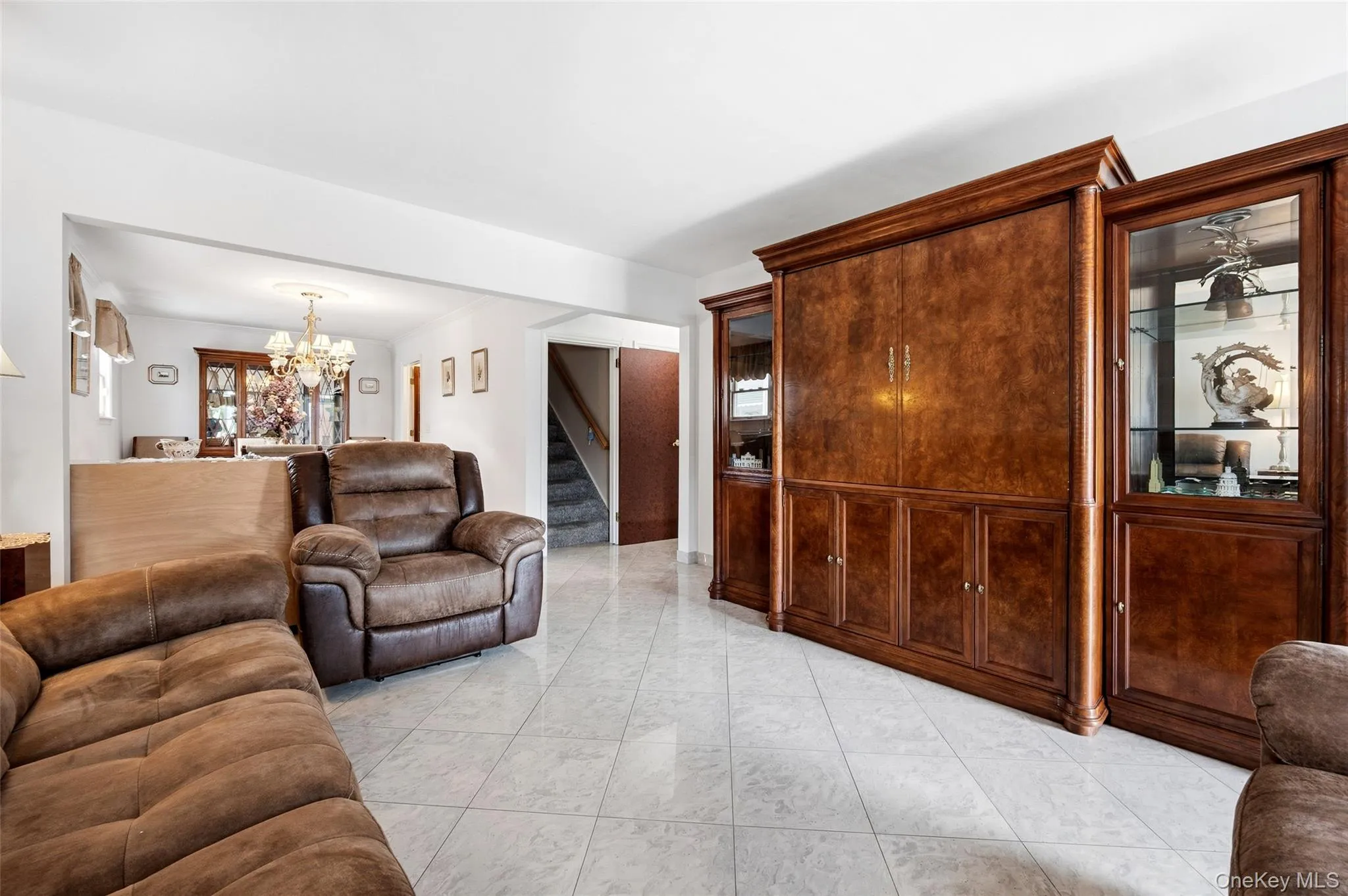 Spacious living area featuring light-toned tile flooring, recessed lighting, and a large built-in wood-finish display unit with glass-front cabinets Spacious living area featuring light-toned tile flooring, recessed lighting, and a large built-in wood-finish display unit with glass-front cabinets