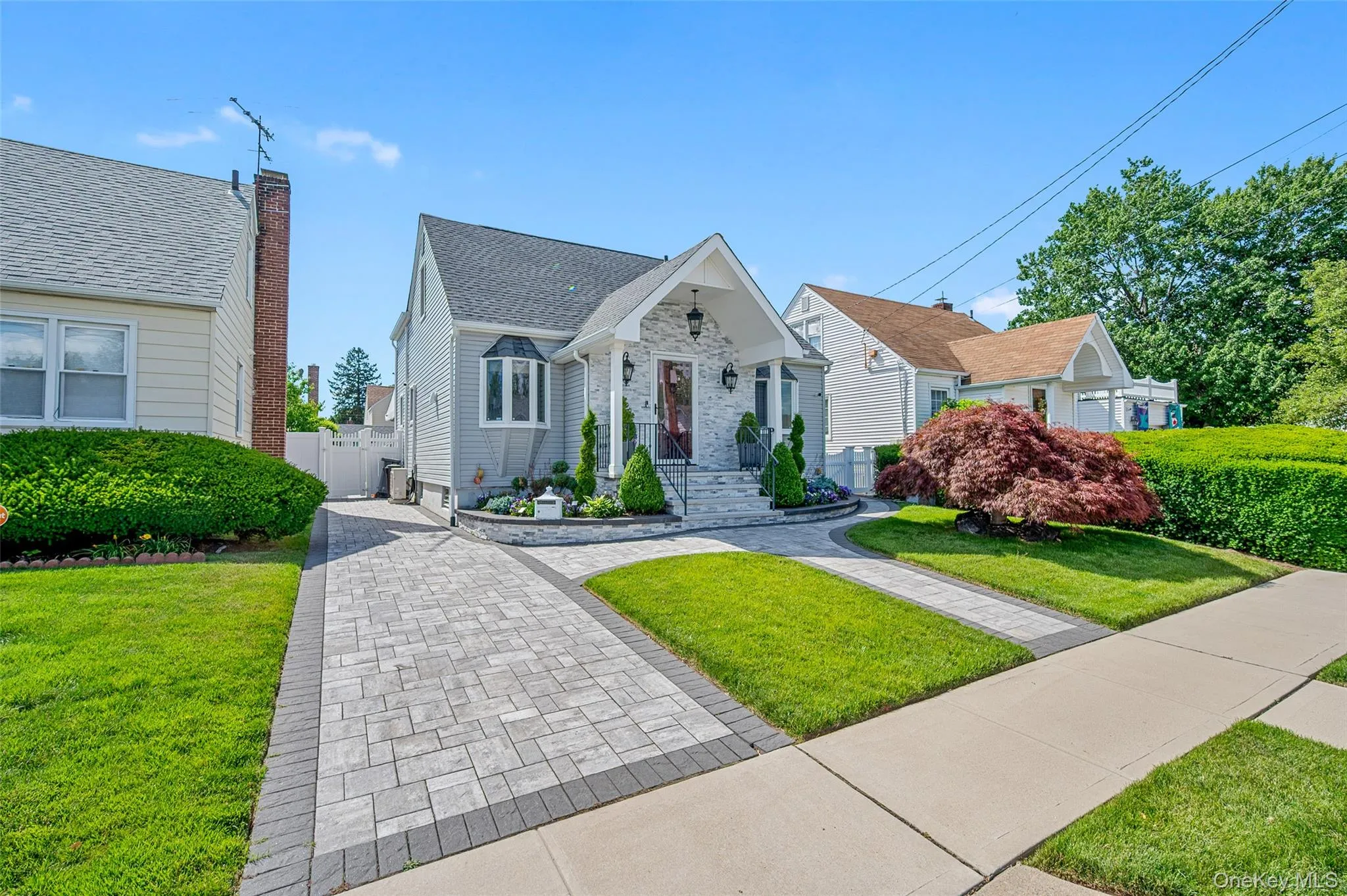 Inviting front exterior featuring a gabled entryway, bay window, light gray siding, and a paver driveway with dark gray border accents Inviting front exterior featuring a gabled entryway, bay window, light gray siding, and a paver driveway with dark gray border accents