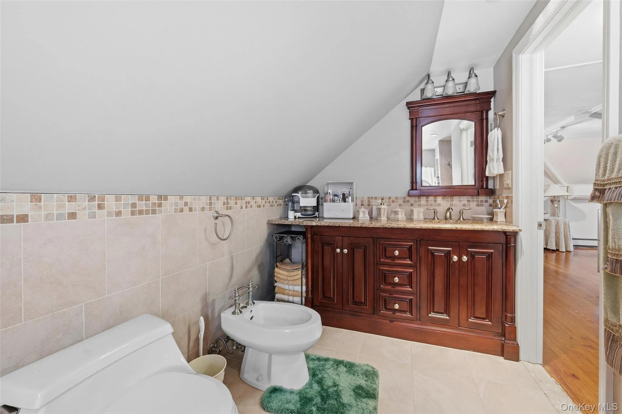 En-suite bathroom featuring a double vanity with a granite-style countertop, a rich wood-finish cabinet, tiled flooring, a tiled accent wall, and a bidet En-suite bathroom featuring a double vanity with a granite-style countertop, a rich wood-finish cabinet, tiled flooring, a tiled accent wall, and a bidet