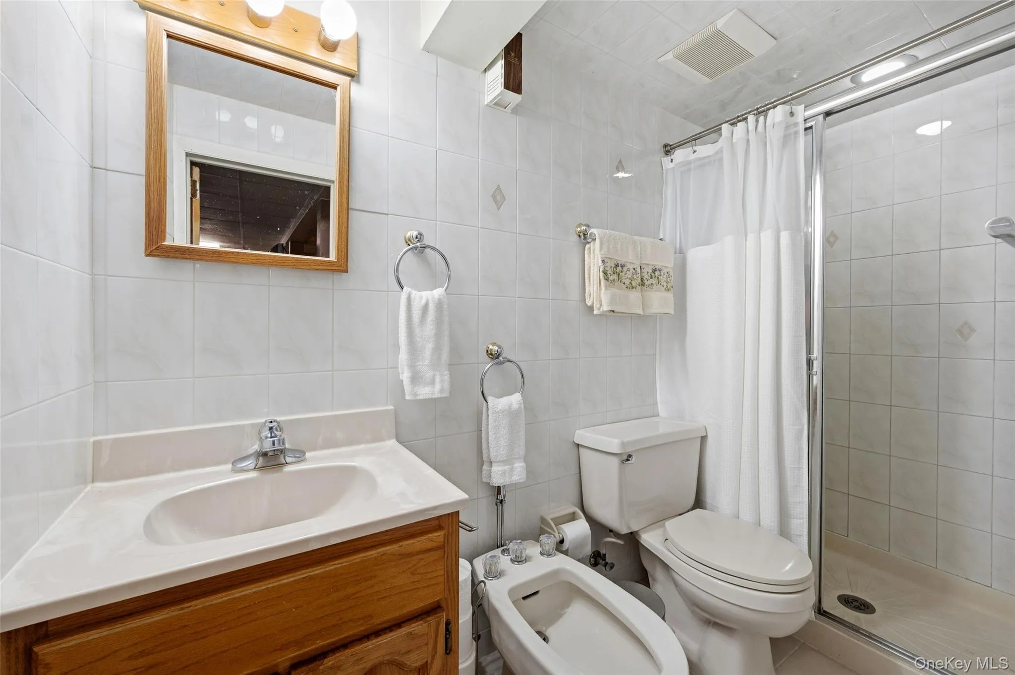Bathroom featuring a light wood vanity with a single basin, a framed mirror with overhead lighting, a toilet, a bidet, and a glass-enclosed shower stall with a white tiled surround Bathroom featuring a light wood vanity with a single basin, a framed mirror with overhead lighting, a toilet, a bidet, and a glass-enclosed shower stall with a white tiled surround