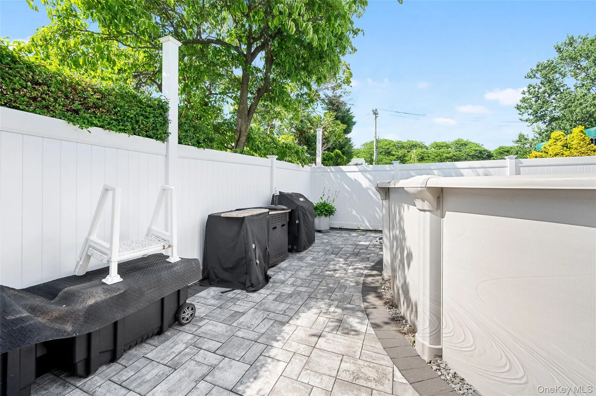 Paved patio area featuring an above-ground pool, a white privacy fence, and mature perimeter trees Paved patio area featuring an above-ground pool, a white privacy fence, and mature perimeter trees