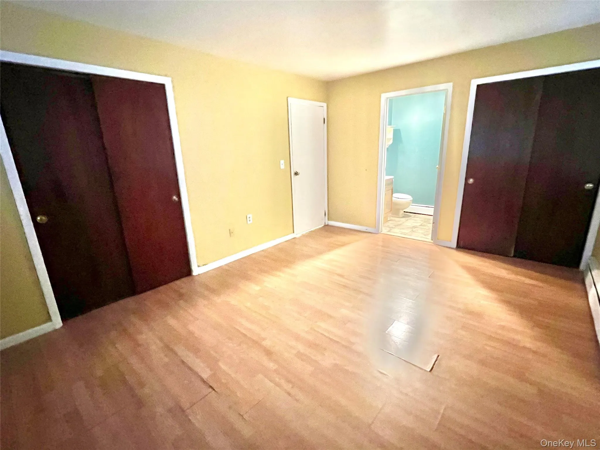 Unfurnished bedroom featuring multiple closets, ensuite bath, wood finished floors, and a baseboard radiator Unfurnished bedroom featuring multiple closets, ensuite bath, wood finished floors, and a baseboard radiator