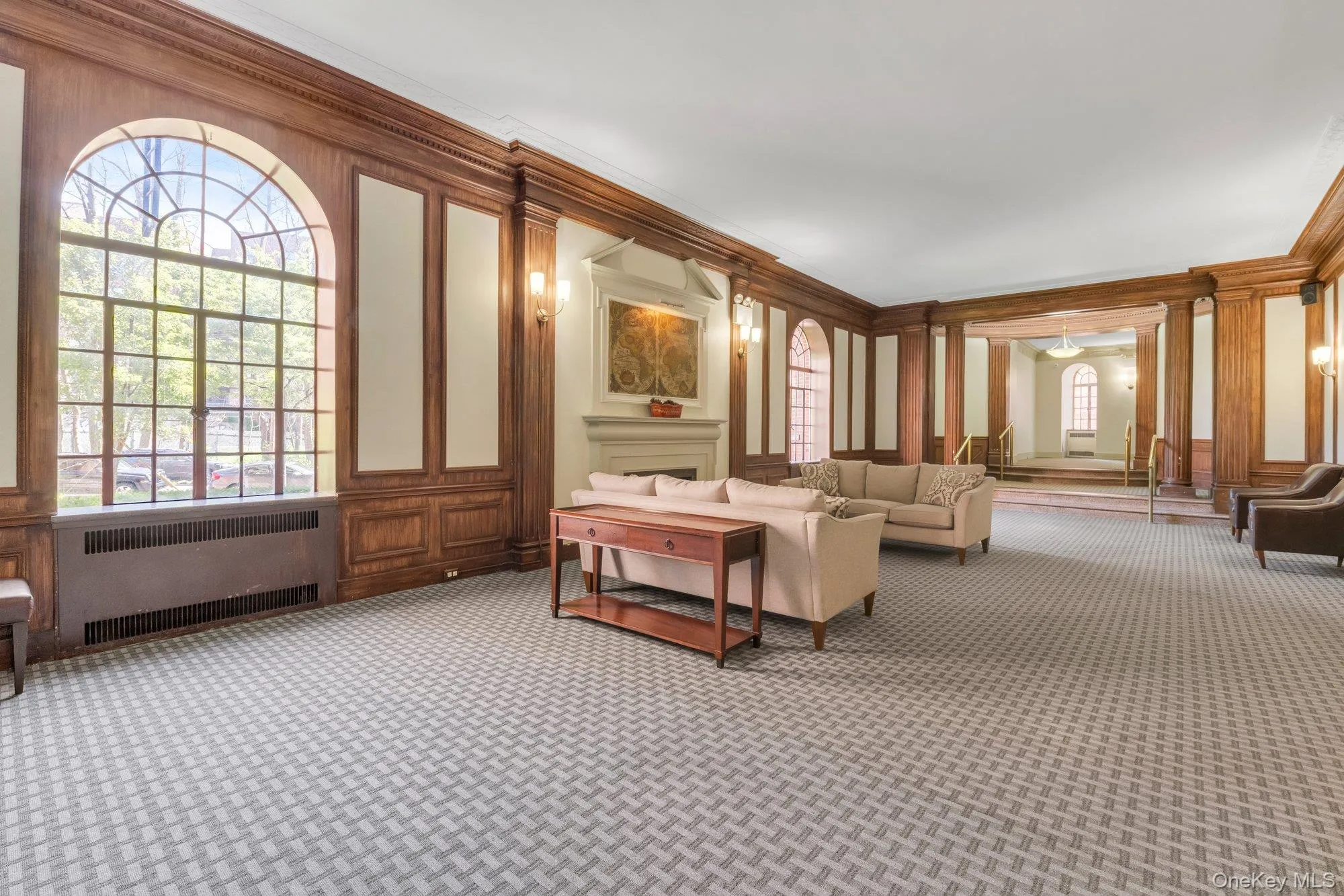 Spacious common area featuring an arched window, wood wall paneling, sconce lighting, a decorative fireplace with mantel, and patterned carpeting Spacious common area featuring an arched window, wood wall paneling, sconce lighting, a decorative fireplace with mantel, and patterned carpeting