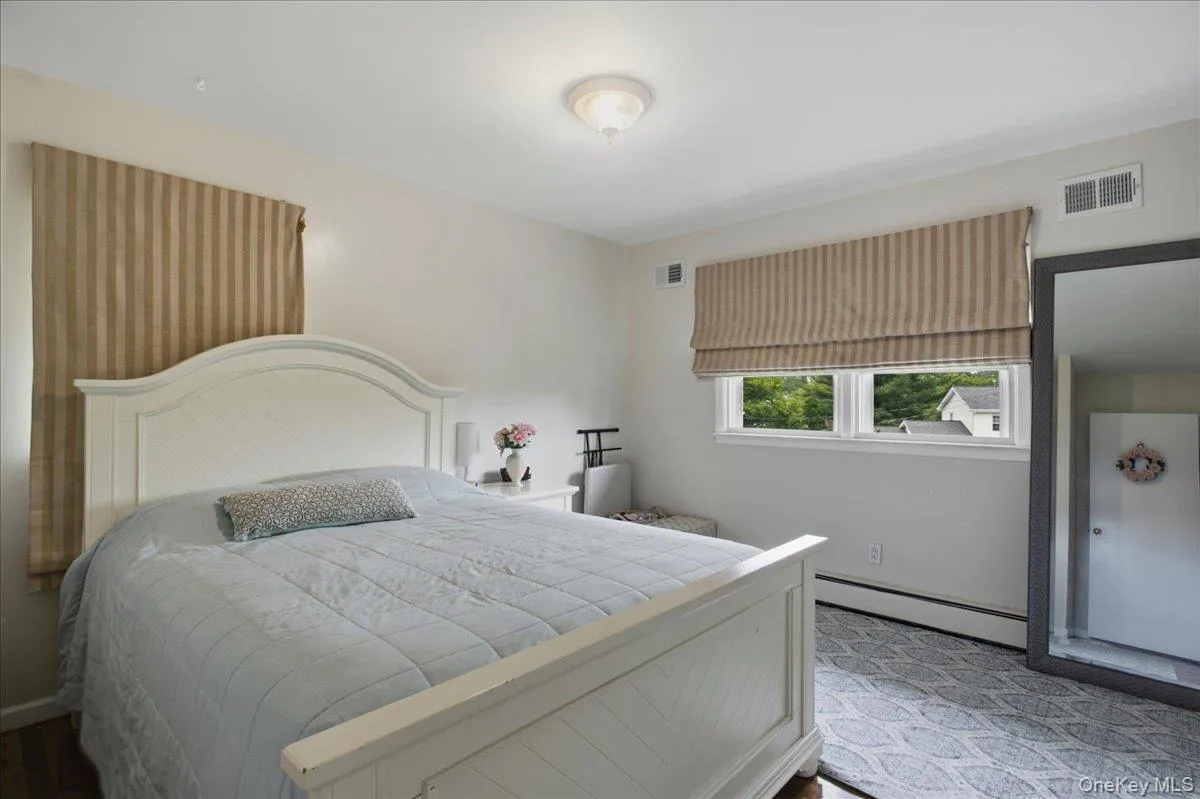 Spacious bedroom featuring light-toned walls, wood-finish flooring, a contemporary ceiling light fixture, and a window with a striped Roman shade Spacious bedroom featuring light-toned walls, wood-finish flooring, a contemporary ceiling light fixture, and a window with a striped Roman shade