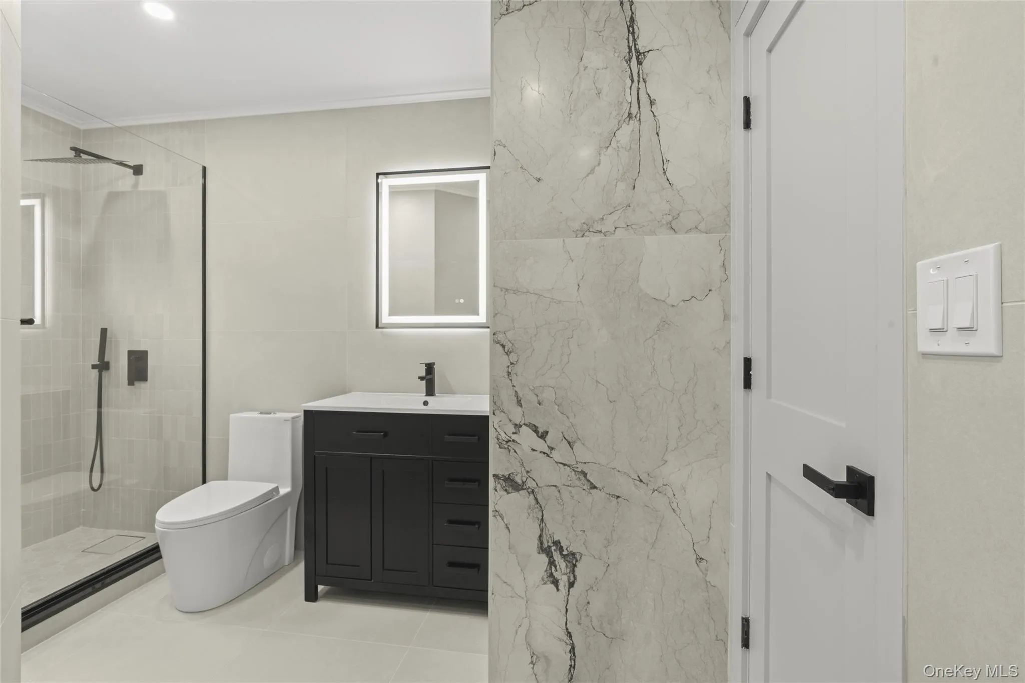 Contemporary bathroom featuring a dark vanity with a white countertop, a lighted mirror, and a frameless glass shower enclosure Contemporary bathroom featuring a dark vanity with a white countertop, a lighted mirror, and a frameless glass shower enclosure