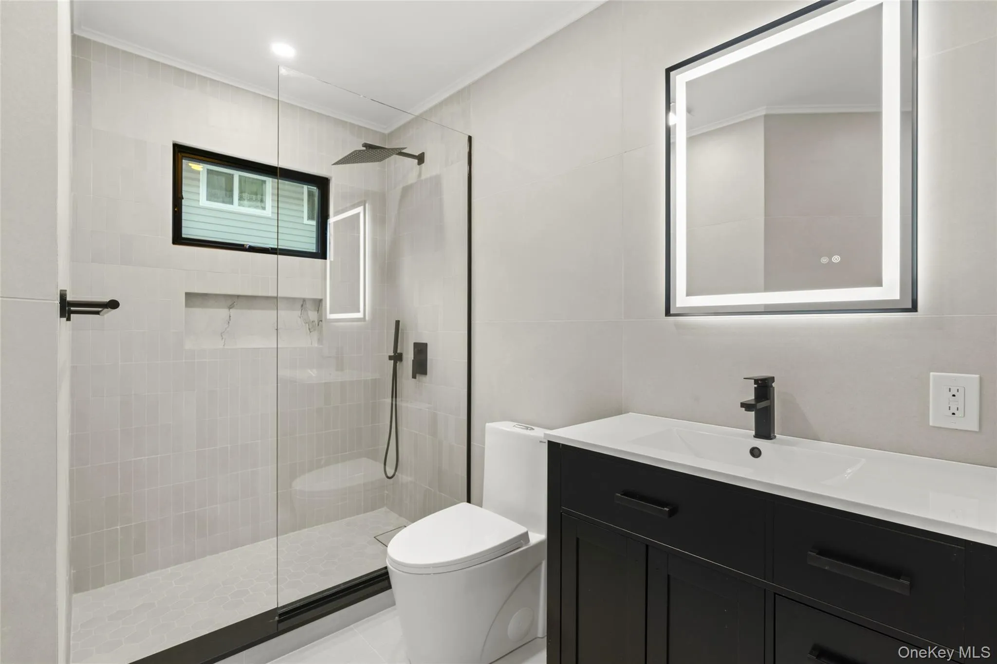 Contemporary bathroom featuring a black framed illuminated mirror, a sleek dark vanity with integrated sink, a glass-enclosed shower with matte black fixtures, a recessed shower niche, and light-toned tile flooring and walls Contemporary bathroom featuring a black framed illuminated mirror, a sleek dark vanity with integrated sink, a glass-enclosed shower with matte black fixtures, a recessed shower niche, and light-toned tile flooring and walls