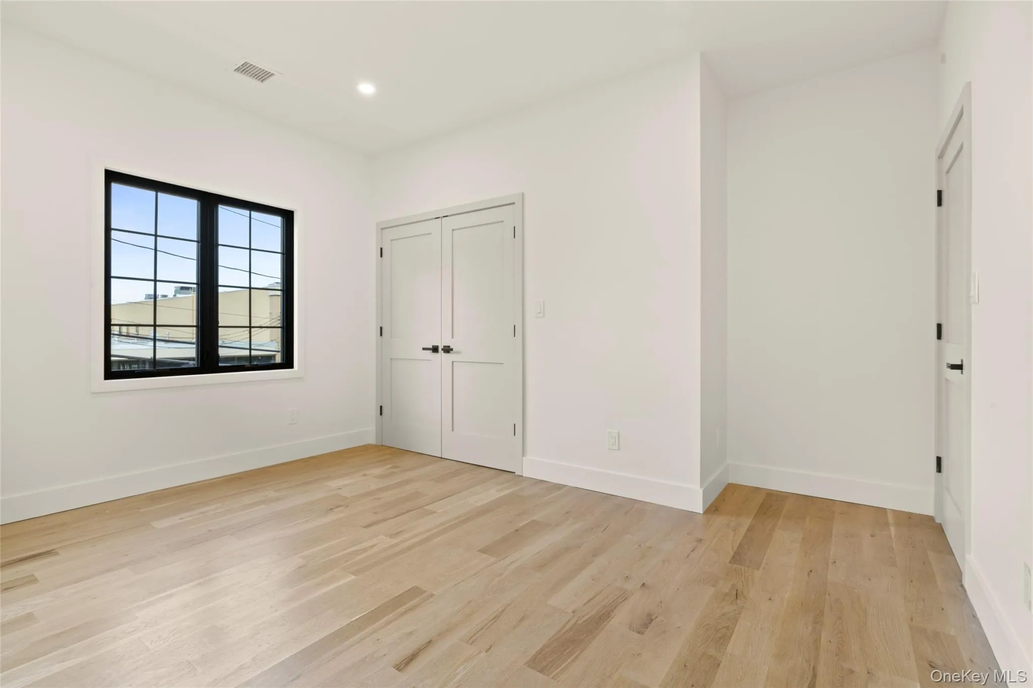 Bright interior space featuring a multi-pane window with black trim, light wood-finish flooring, white walls, recessed ceiling lighting, and white double doors Bright interior space featuring a multi-pane window with black trim, light wood-finish flooring, white walls, recessed ceiling lighting, and white double doors