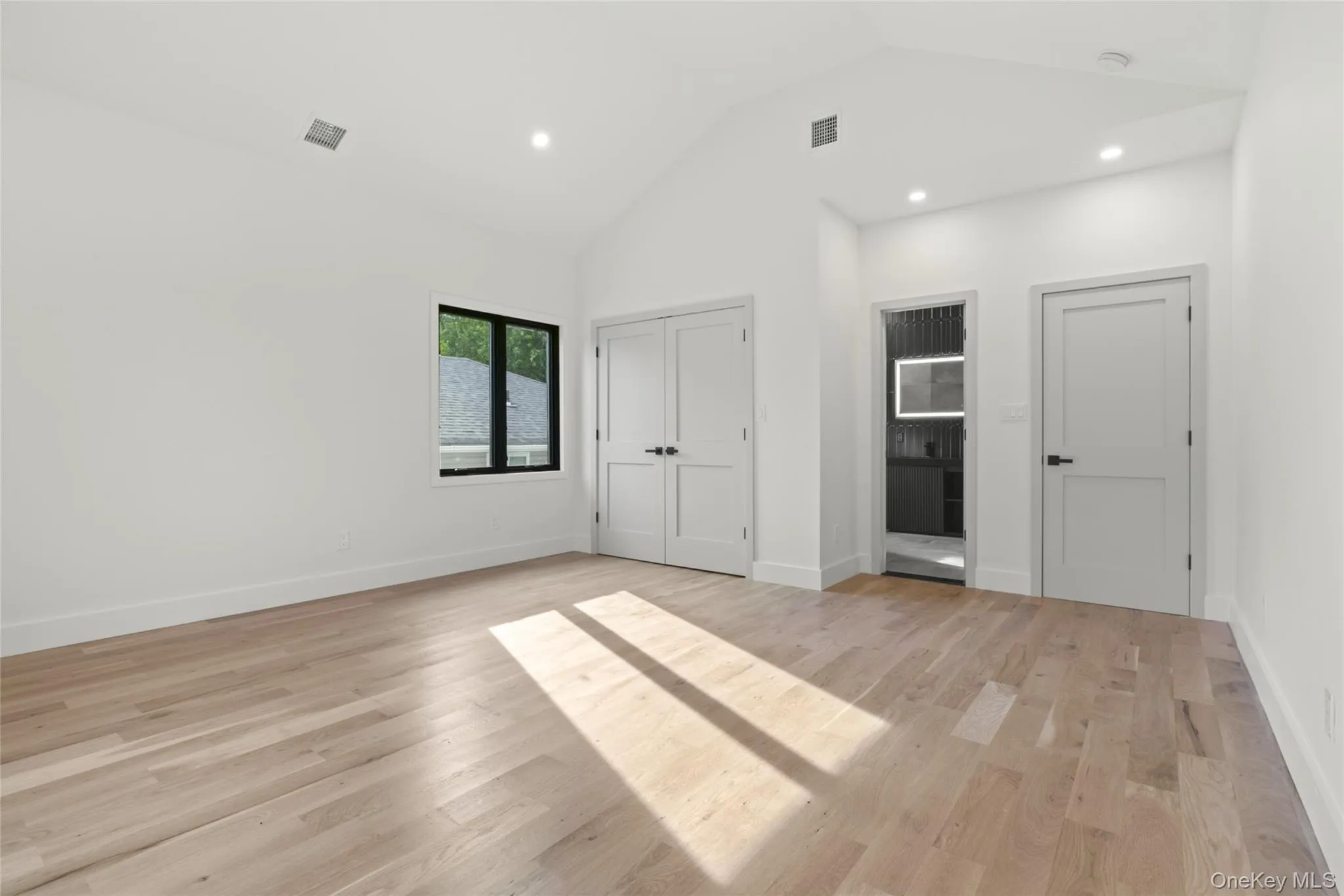 Spacious room featuring vaulted ceilings, light wood-finish flooring, recessed lighting, and a black-framed window Spacious room featuring vaulted ceilings, light wood-finish flooring, recessed lighting, and a black-framed window