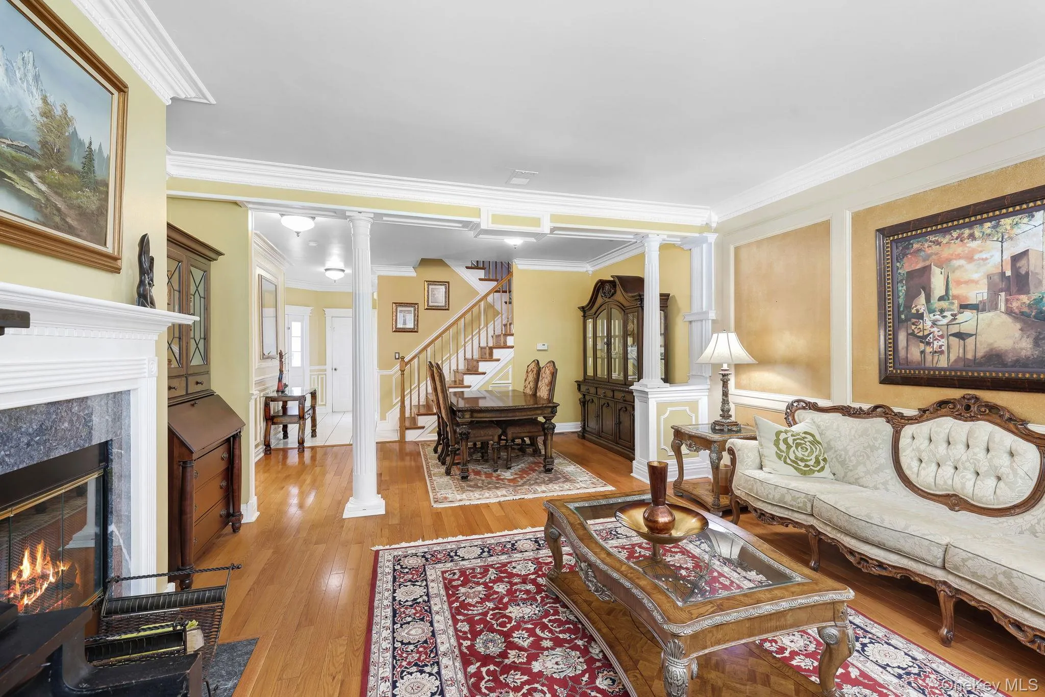 Open-concept living area featuring wood-finish flooring, a white mantel fireplace with a dark stone surround, decorative columns, and crown molding Open-concept living area featuring wood-finish flooring, a white mantel fireplace with a dark stone surround, decorative columns, and crown molding