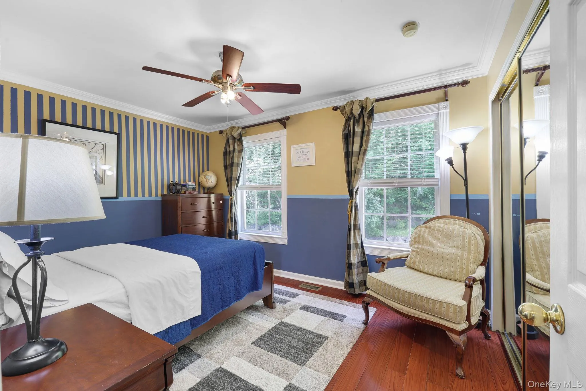 Bright bedroom featuring dual windows, hardwood-finish flooring, a ceiling fan with light, crown molding, and a mirrored closet door Bright bedroom featuring dual windows, hardwood-finish flooring, a ceiling fan with light, crown molding, and a mirrored closet door
