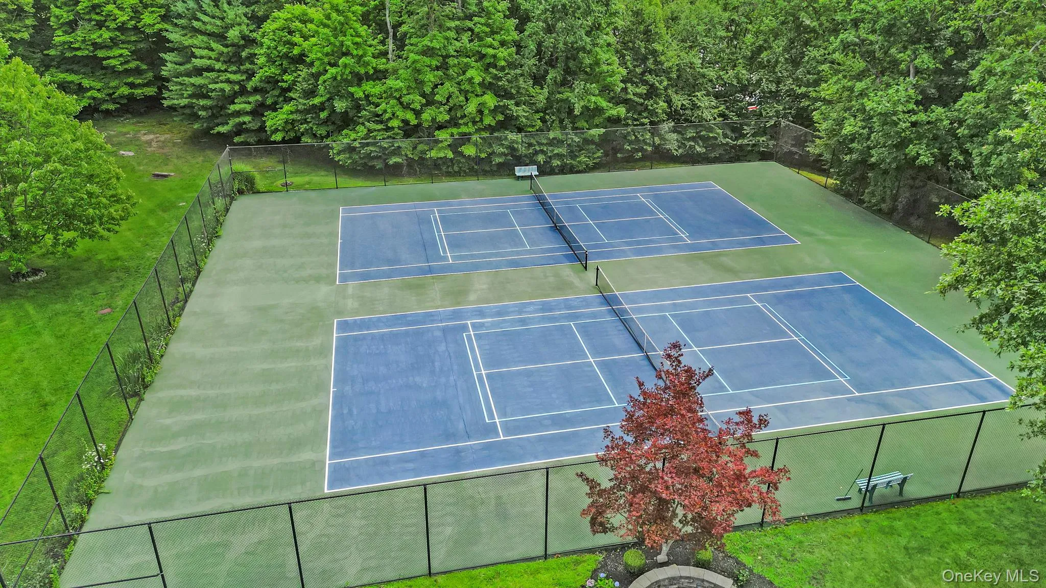 Two full-size fenced tennis courts featuring blue playing surfaces and green surrounds Two full-size fenced tennis courts featuring blue playing surfaces and green surrounds