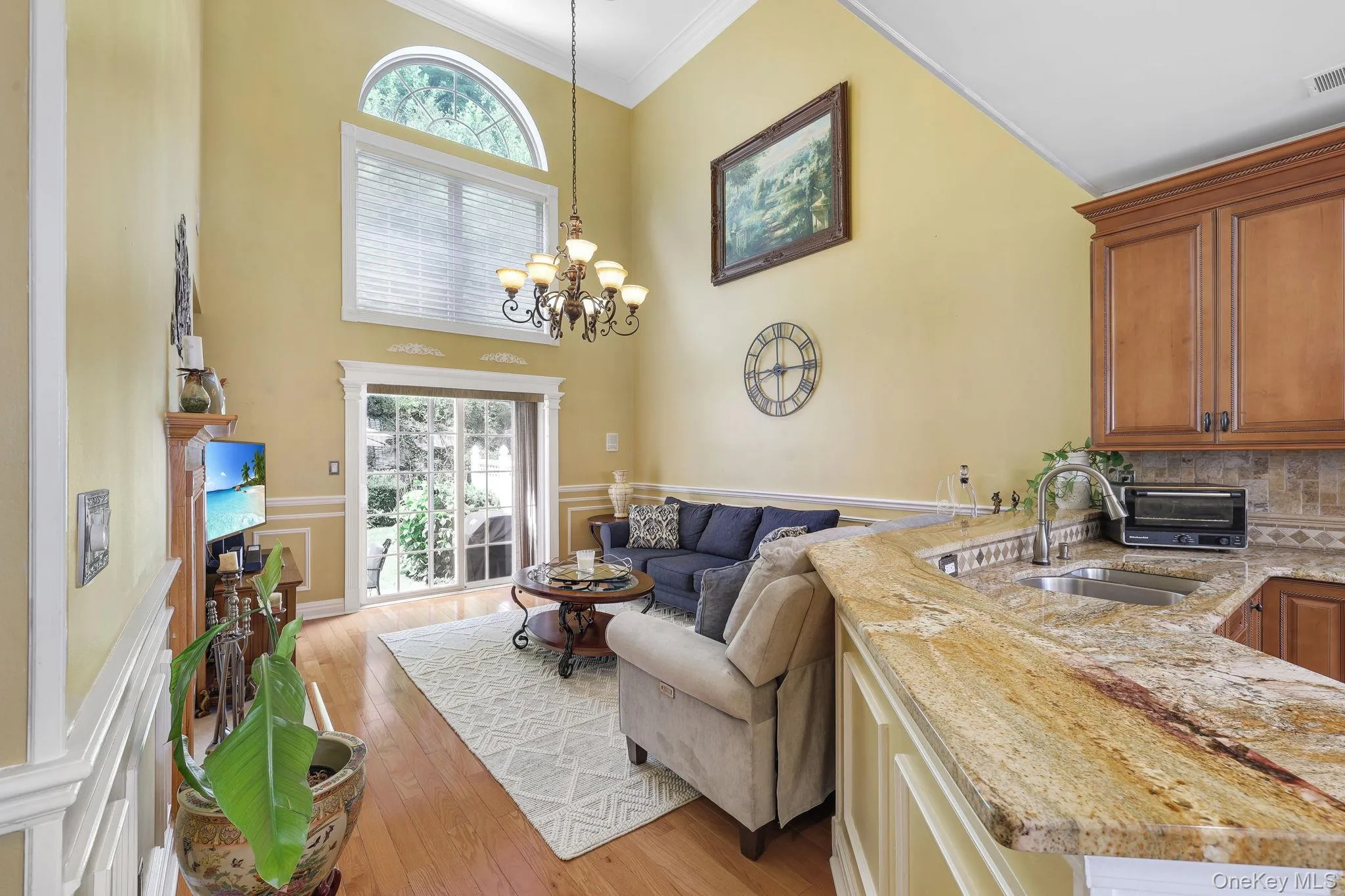 Open-concept living space featuring high ceilings, wood-finish flooring, and a two-story window with an arched transom access to outdoor patio and backyard Open-concept living space featuring high ceilings, wood-finish flooring, and a two-story window with an arched transom access to outdoor patio and backyard