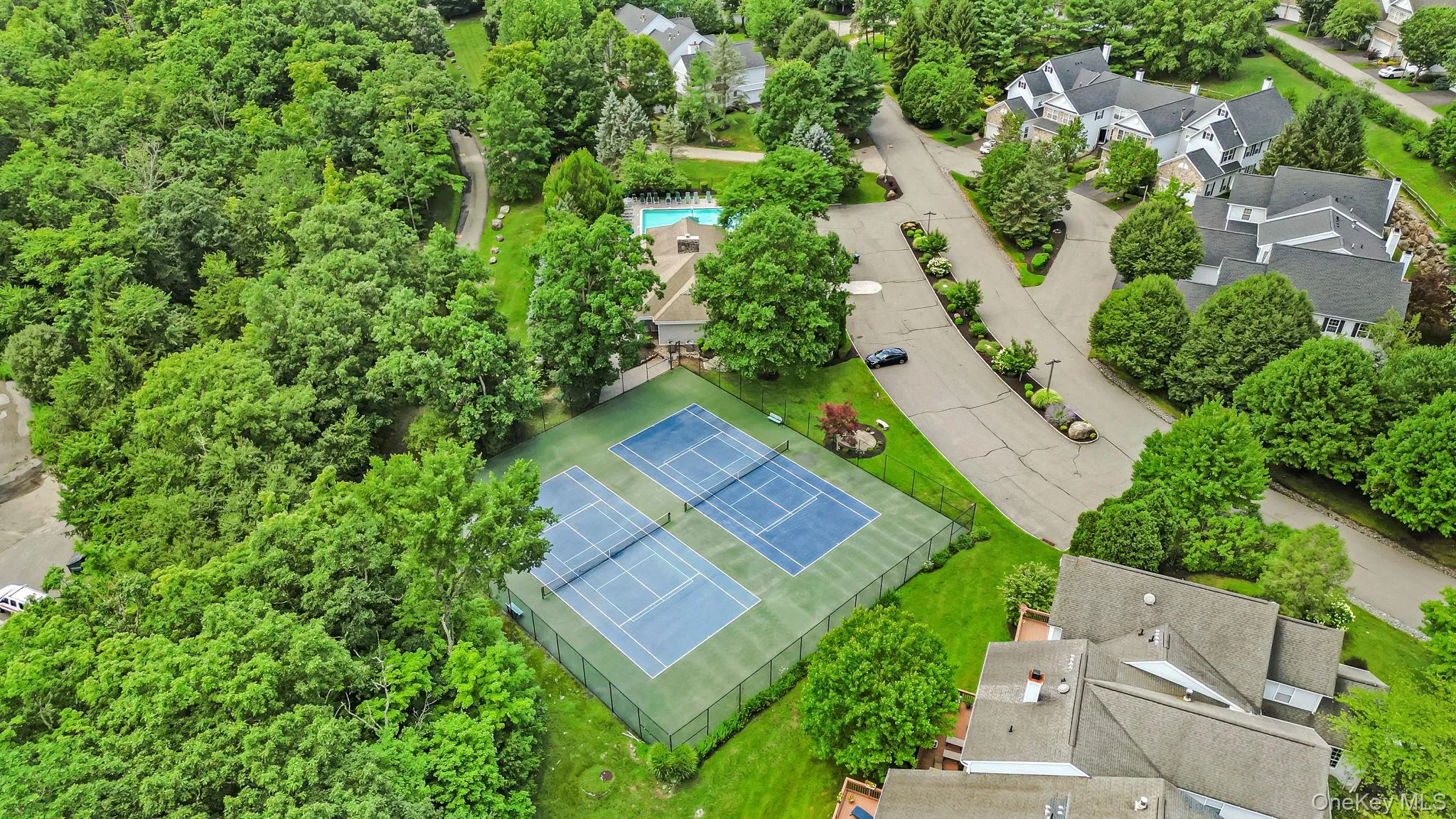 Aerial perspective showcasing dual tennis courts with a dark perimeter fence, a nearby swimming pool with a paved surround, and surrounding mature trees Aerial perspective showcasing dual tennis courts with a dark perimeter fence, a nearby swimming pool with a paved surround, and surrounding mature trees
