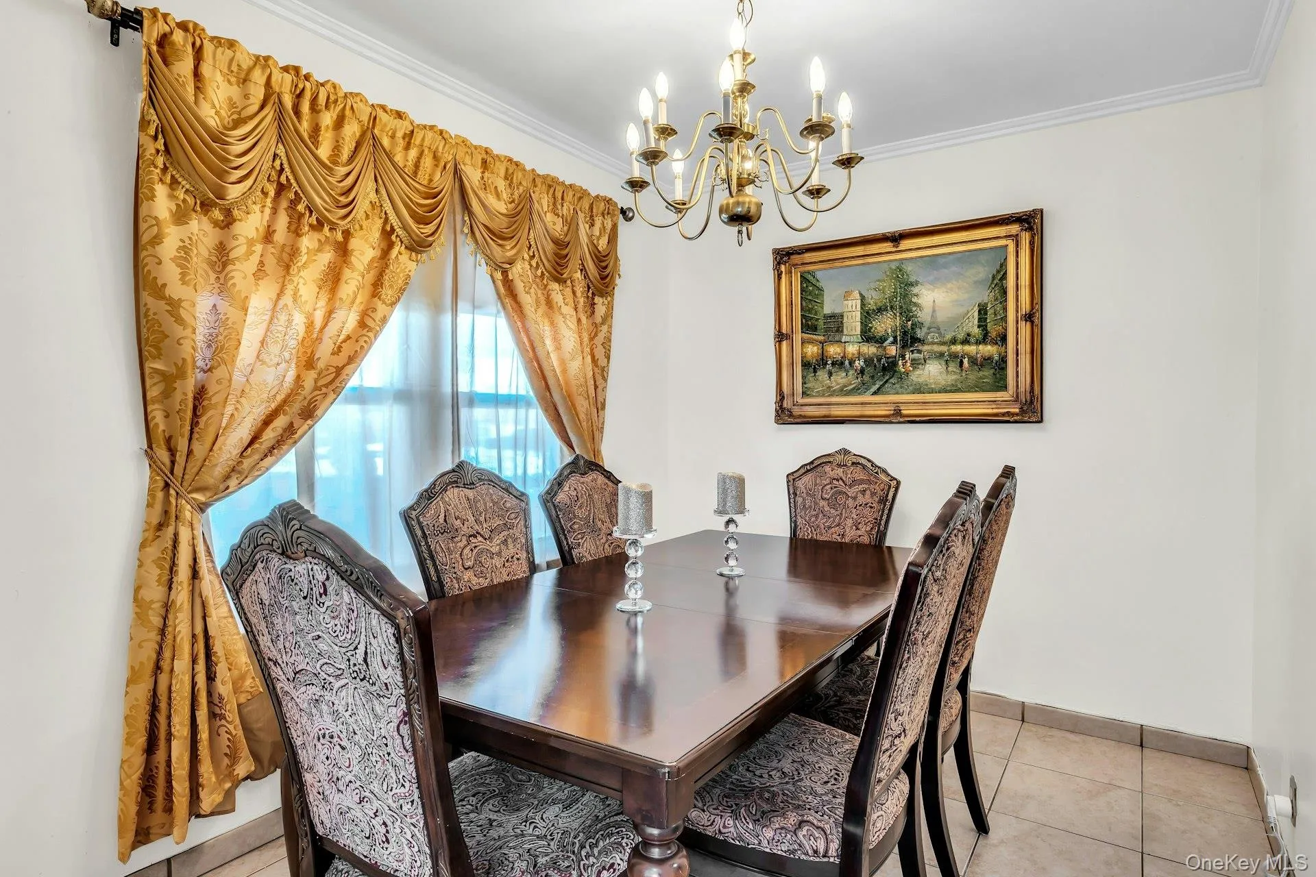 Dining area featuring a dark wood-finish table with upholstered chairs, a brass chandelier, crown molding, and tiled flooring Dining area featuring a dark wood-finish table with upholstered chairs, a brass chandelier, crown molding, and tiled flooring