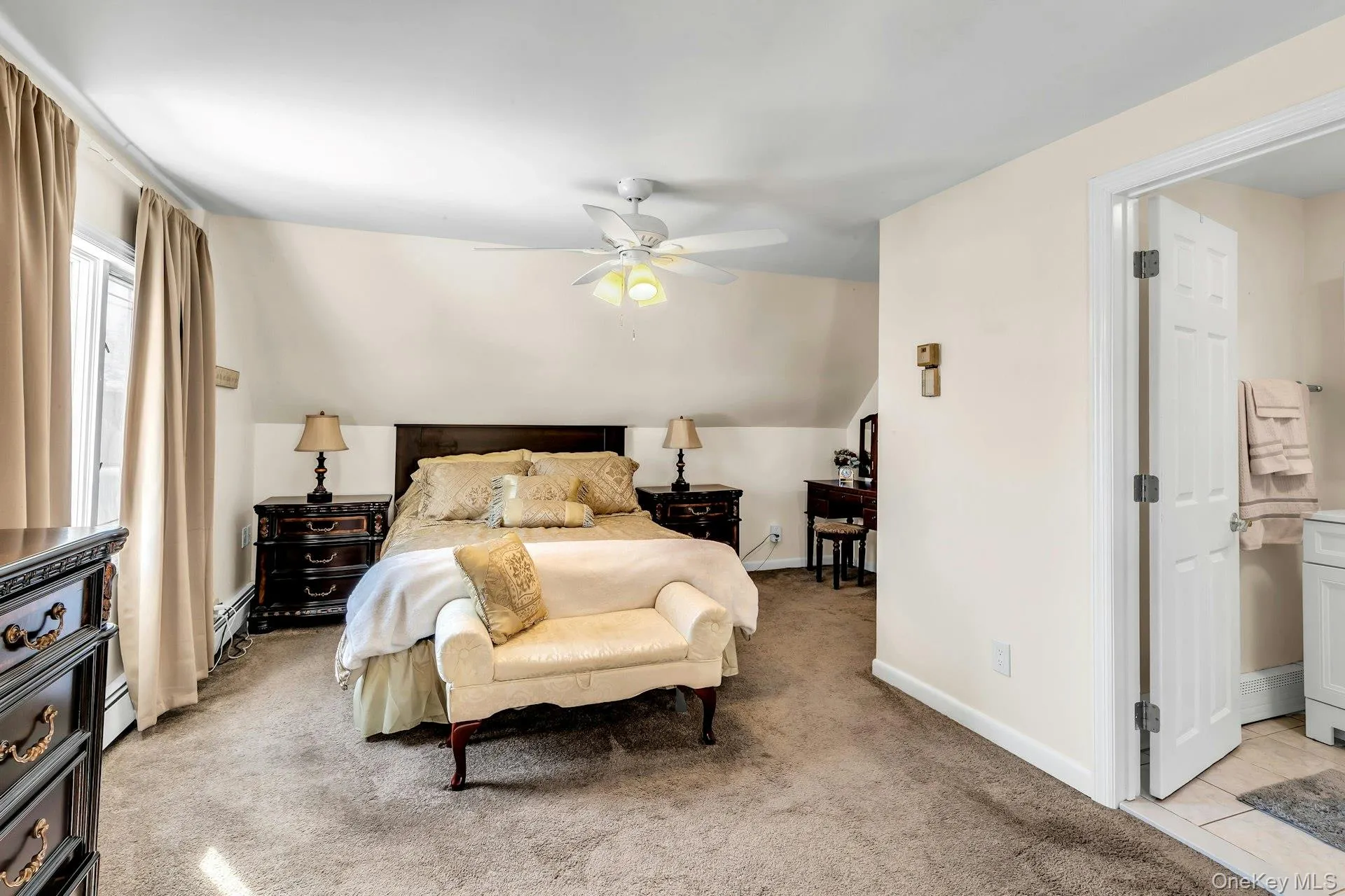 Carpeted bedroom featuring a ceiling fan with integrated lighting, a window with natural light, and an open doorway revealing a tiled bathroom Carpeted bedroom featuring a ceiling fan with integrated lighting, a window with natural light, and an open doorway revealing a tiled bathroom