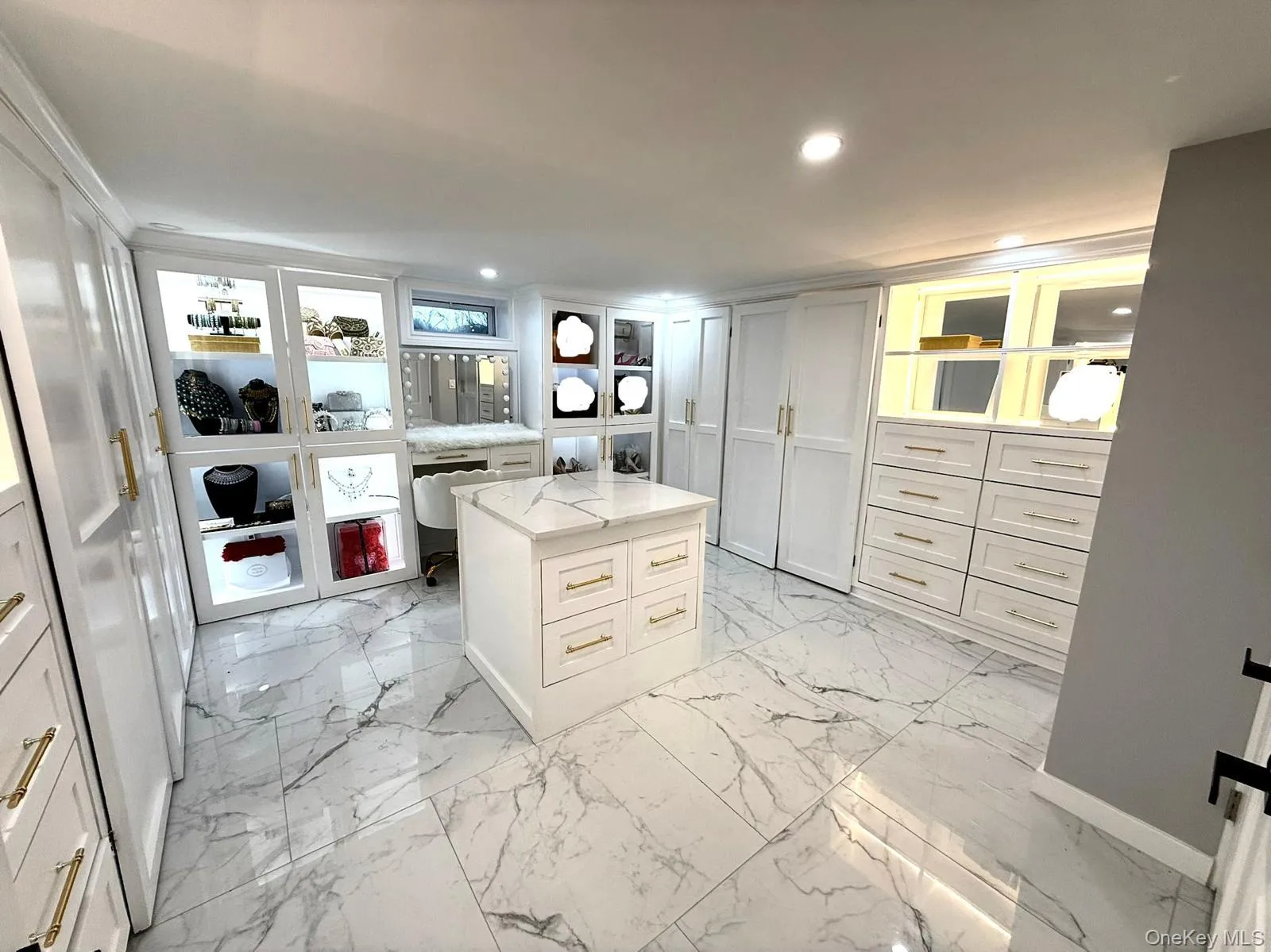 Custom closet system featuring a central island with a stone-finish countertop, integrated recessed lighting, extensive built-in cabinetry with gold-tone hardware, display shelving with backlighting, and large-format porcelain tile flooring Custom closet system featuring a central island with a stone-finish countertop, integrated recessed lighting, extensive built-in cabinetry with gold-tone hardware, display shelving with backlighting, and large-format porcelain tile flooring
