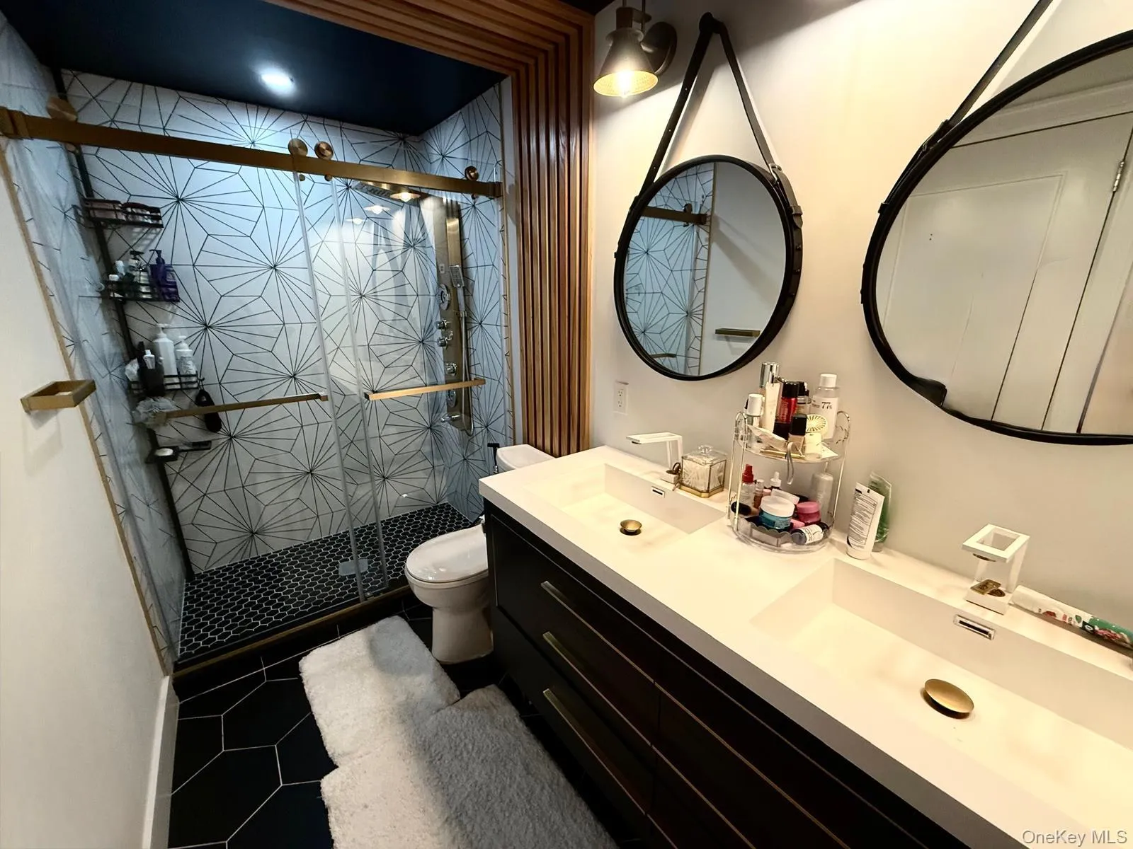 Modern bathroom featuring a dual vanity with integrated sinks, a glass-enclosed shower with geometric tile surround, black hexagonal floor tiles, and wood slat wall detailing Modern bathroom featuring a dual vanity with integrated sinks, a glass-enclosed shower with geometric tile surround, black hexagonal floor tiles, and wood slat wall detailing