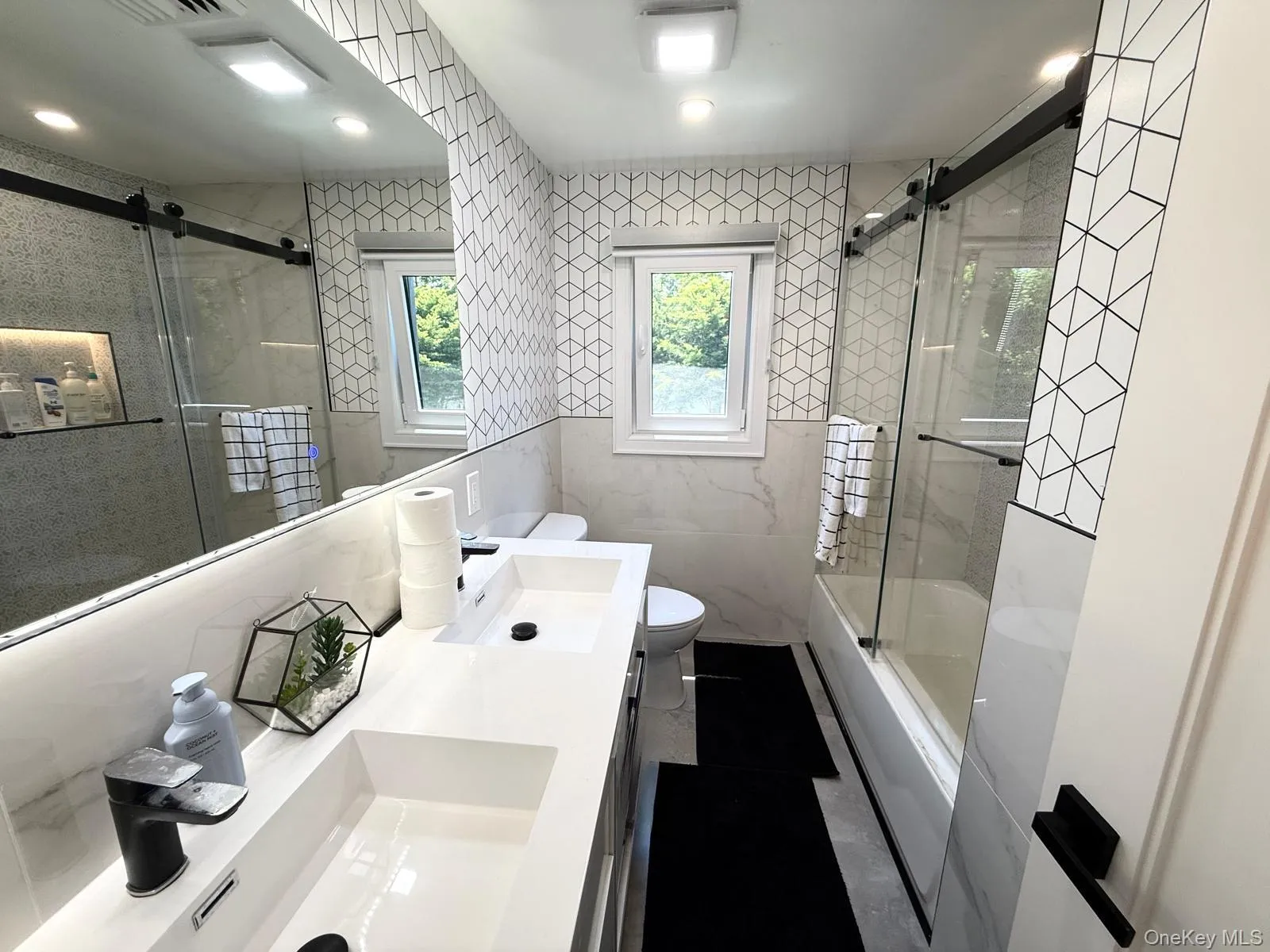 Bathroom featuring a dual vanity with integrated sinks, black finish faucets, and a full-width mirror with integrated lighting Bathroom featuring a dual vanity with integrated sinks, black finish faucets, and a full-width mirror with integrated lighting