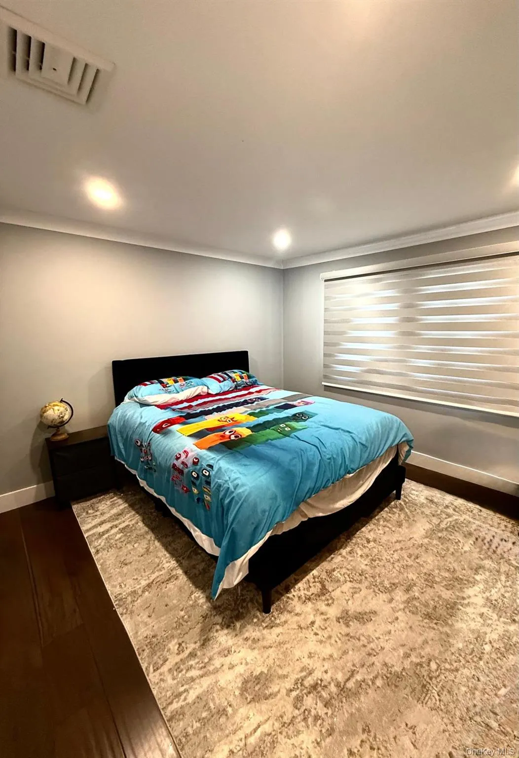 Recessed lighting illuminates this room featuring dark wood-finish flooring, a patterned area rug, and a window with horizontal blinds Recessed lighting illuminates this room featuring dark wood-finish flooring, a patterned area rug, and a window with horizontal blinds
