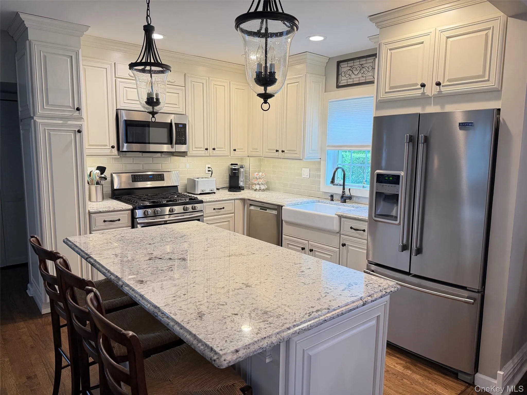 Kitchen featuring a large island with a stone countertop, white shaker-style cabinetry, stainless steel appliances, a white farmhouse sink, and wood-finish flooring Kitchen featuring a large island with a stone countertop, white shaker-style cabinetry, stainless steel appliances, a white farmhouse sink, and wood-finish flooring