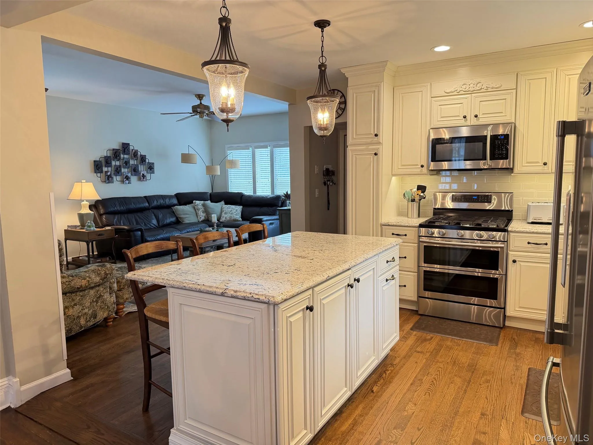 Kitchen featuring a granite-topped island with seating, stainless steel appliances, white cabinetry with decorative molding, pendant lighting, and wood-finish flooring Kitchen featuring a granite-topped island with seating, stainless steel appliances, white cabinetry with decorative molding, pendant lighting, and wood-finish flooring