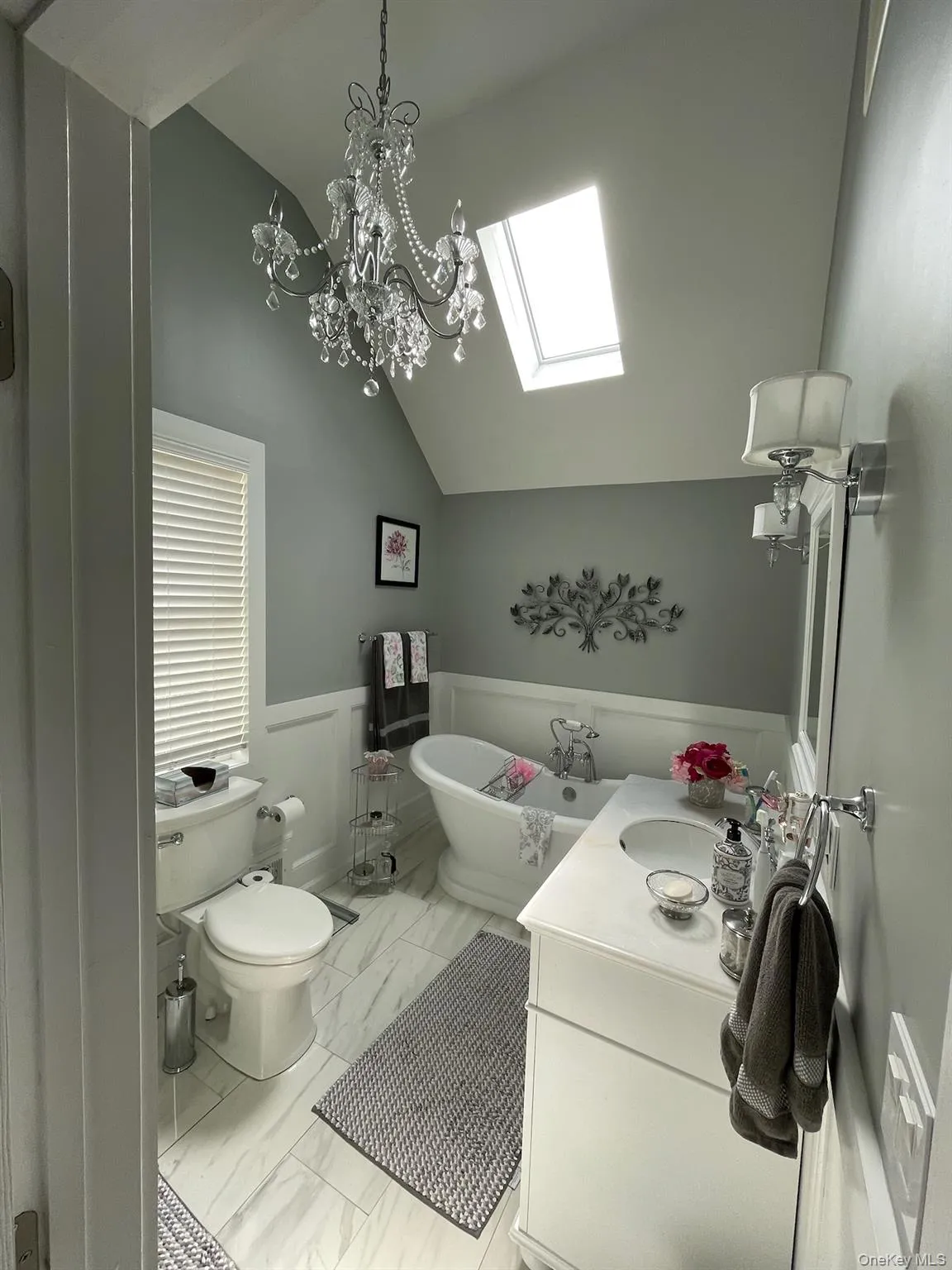 Bathroom featuring a freestanding pedestal tub, a crystal chandelier, a white vanity with an integrated sink, a skylight, and large format tile flooring Bathroom featuring a freestanding pedestal tub, a crystal chandelier, a white vanity with an integrated sink, a skylight, and large format tile flooring