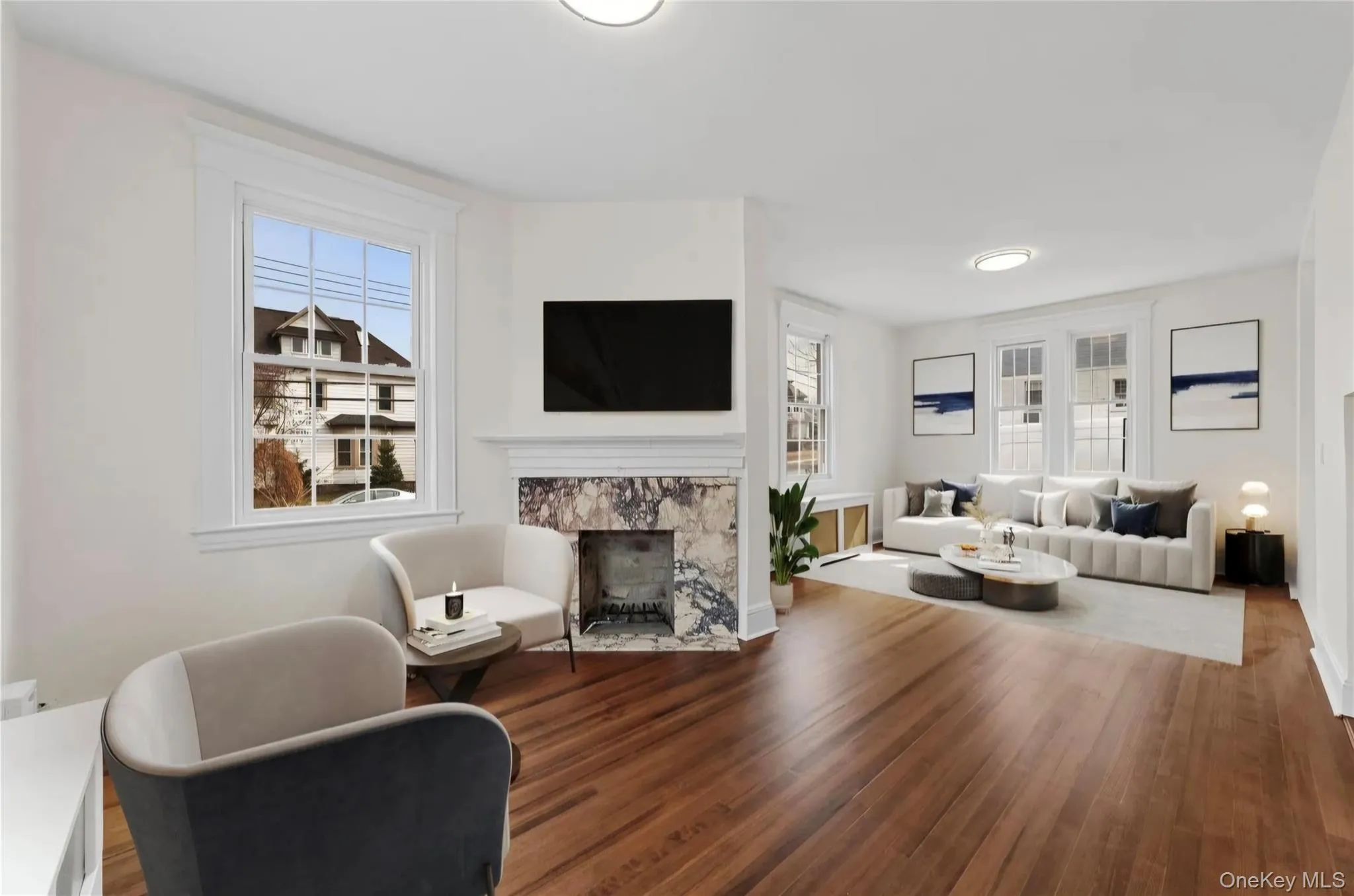 Spacious room featuring rich wood-finish flooring, a white mantel fireplace with a marble-style surround, and multiple large windows Spacious room featuring rich wood-finish flooring, a white mantel fireplace with a marble-style surround, and multiple large windows